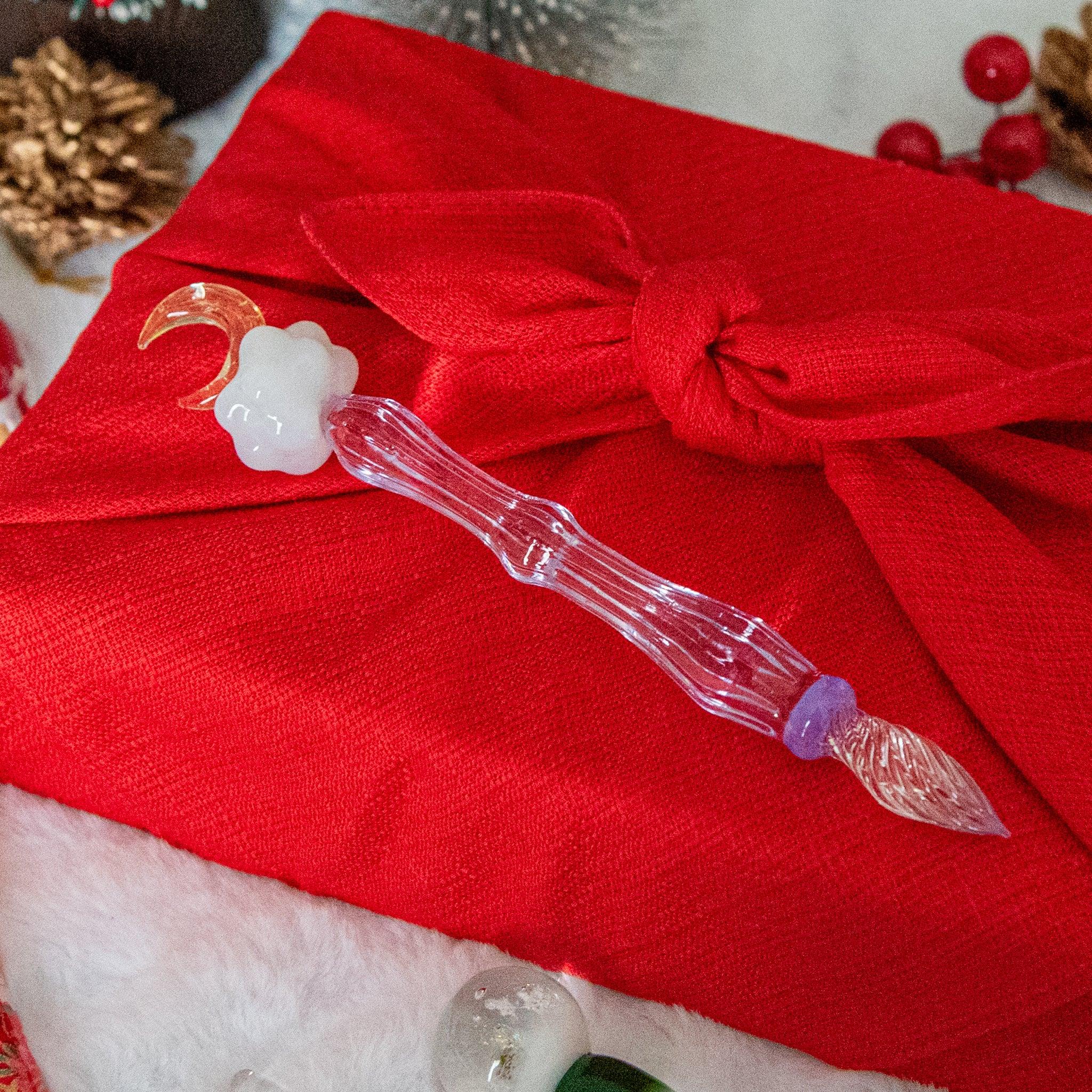 A glass pen with a moon-shaped ornament and a fluffy cloud design lies on a red cloth-wrapped gift, surrounded by festive holiday decor.