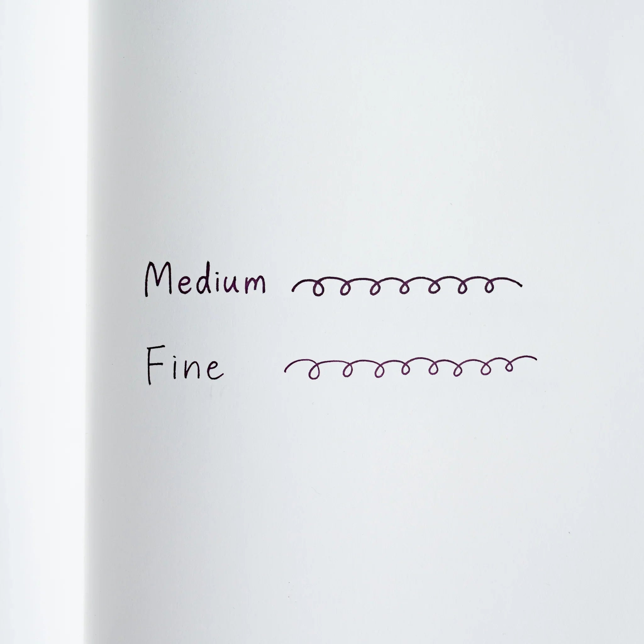 Ink writing sample on white paper, looped cursive lines labeled Medium and Fine showing the two line widths in purple ink