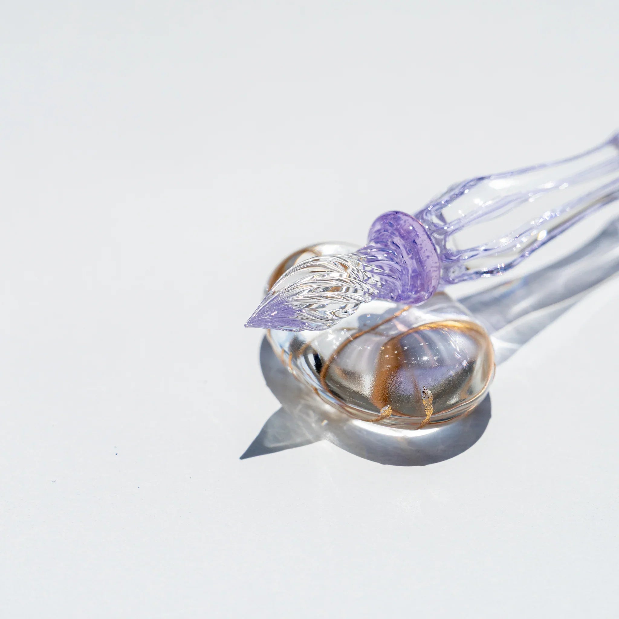 Angled view of the Moonlit Cloud glass dip pen nib, clear grooved tip with lavender collar, on a gold flecked glass rest