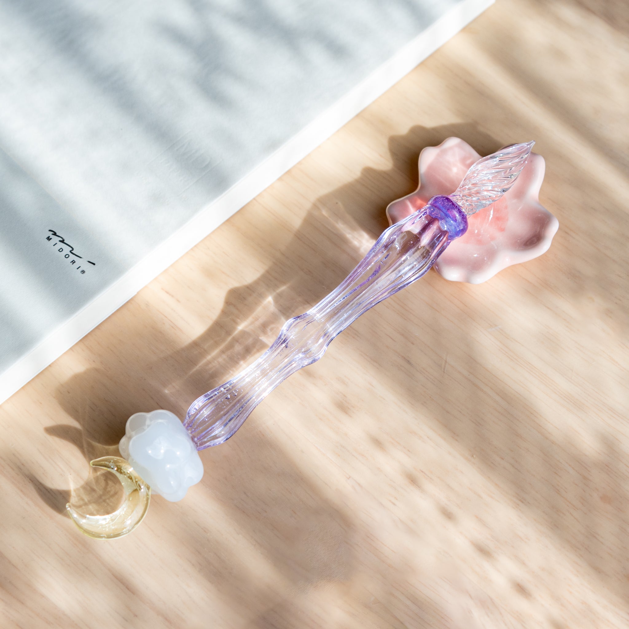 A delicate glass pen with a moon and cloud motif, resting on a wooden surface with soft shadows. The pen’s transparent lavender body beautifully reflects light, showcasing its craftsmanship.