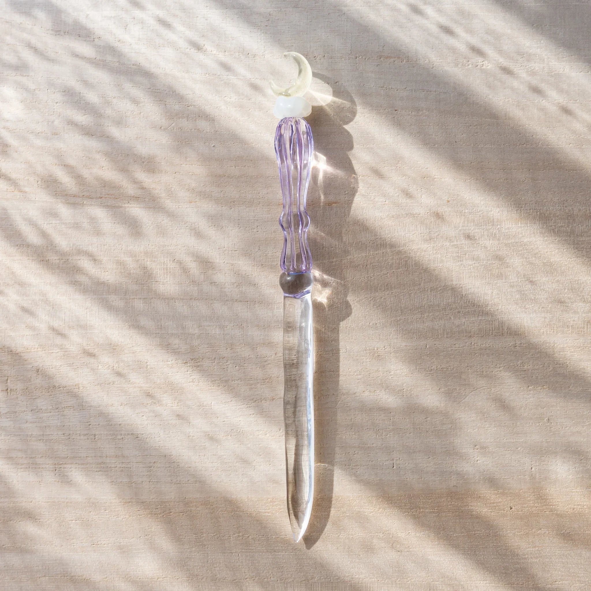 Lavender-colored glass letter opener with a moon and cloud motif placed vertically on a sunlit desk, casting soft shadows.