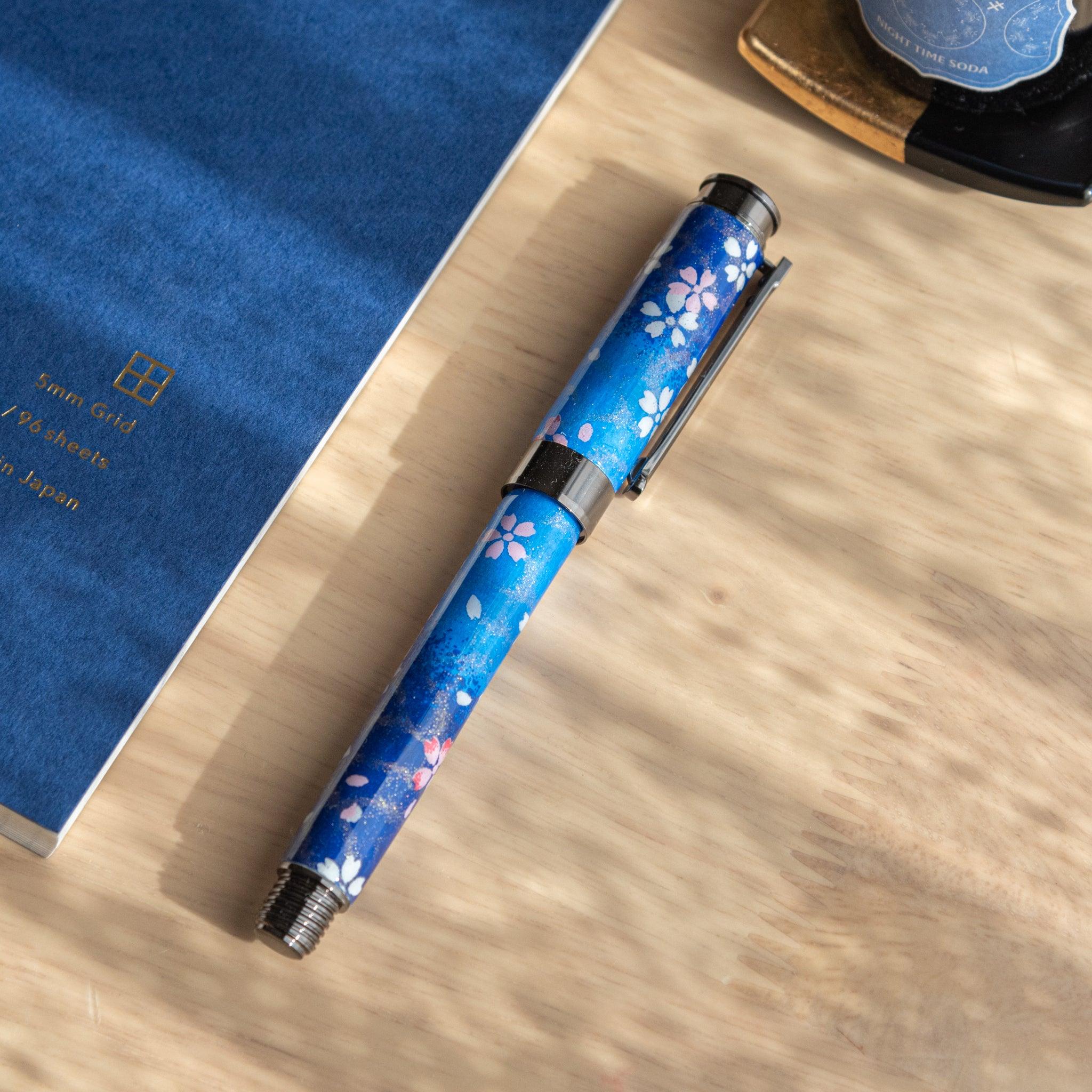 Closed Japanese fountain pen with moonlit sakura design in blue tones, placed beside a matching blue notebook. (98 characters)