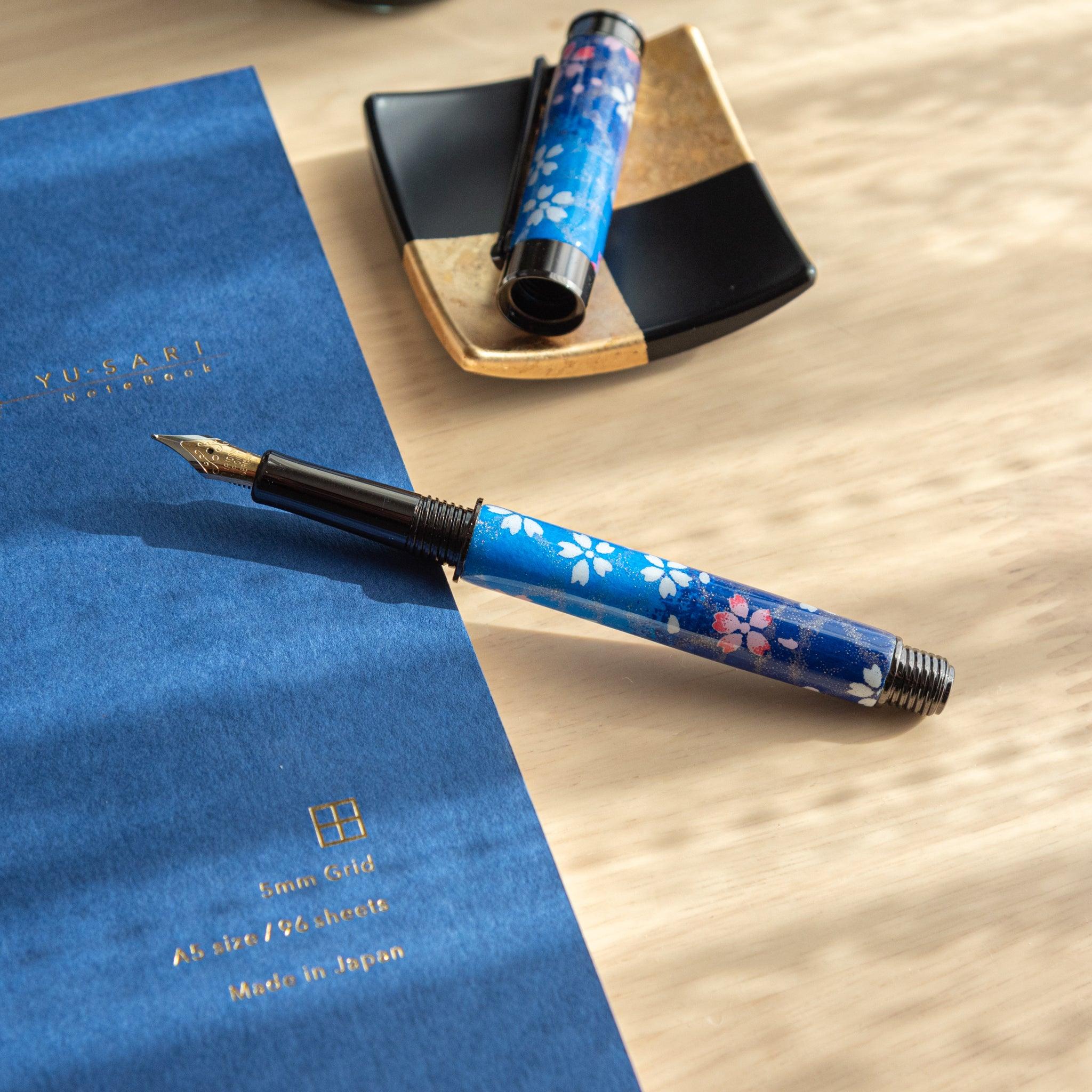 Open Japanese fountain pen featuring a blue sakura design, displayed on a blue notebook and gold-accented tray. (94 characters)