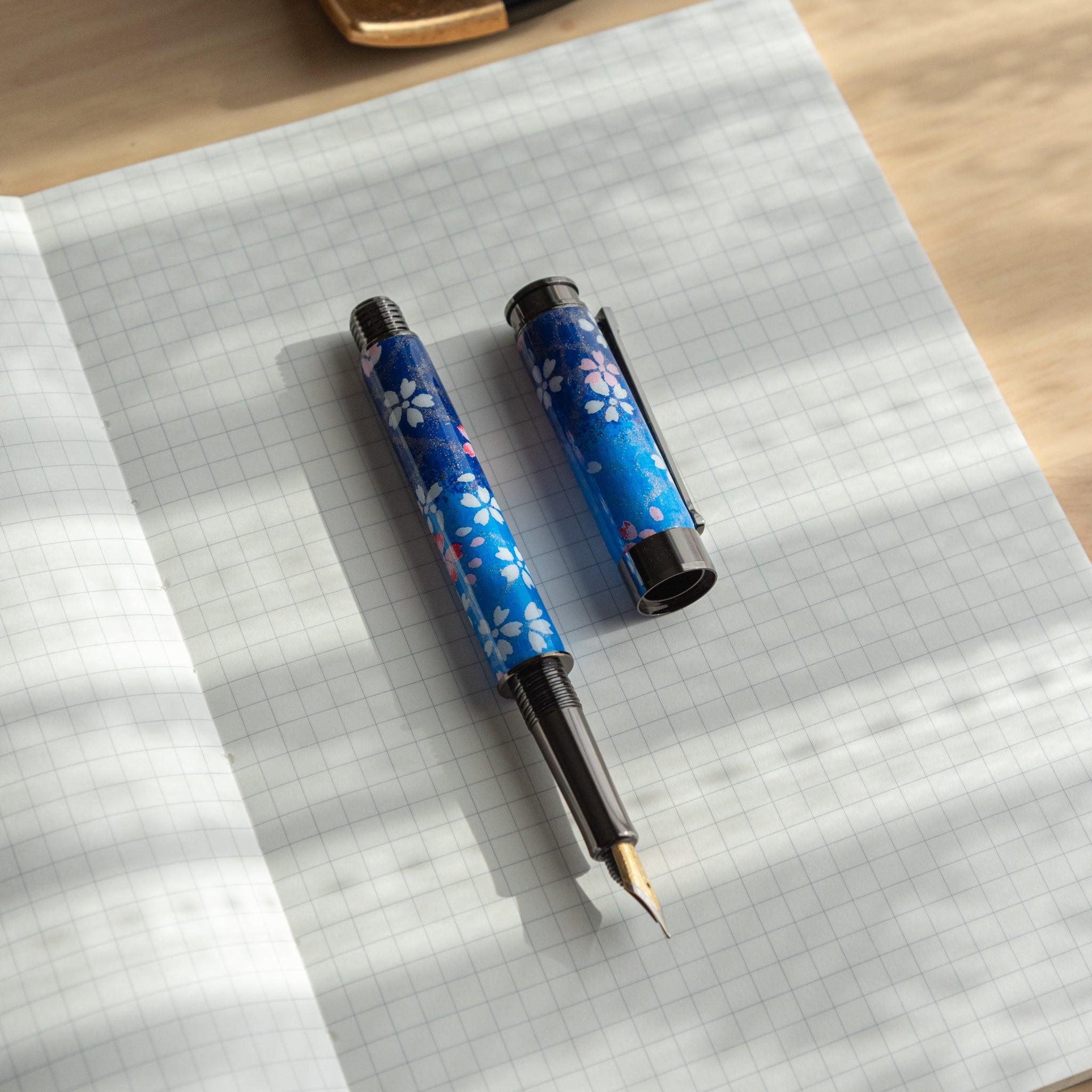 Open Japanese fountain pen with blue sakura design, placed on a grid-lined notebook under natural light. (91 characters)