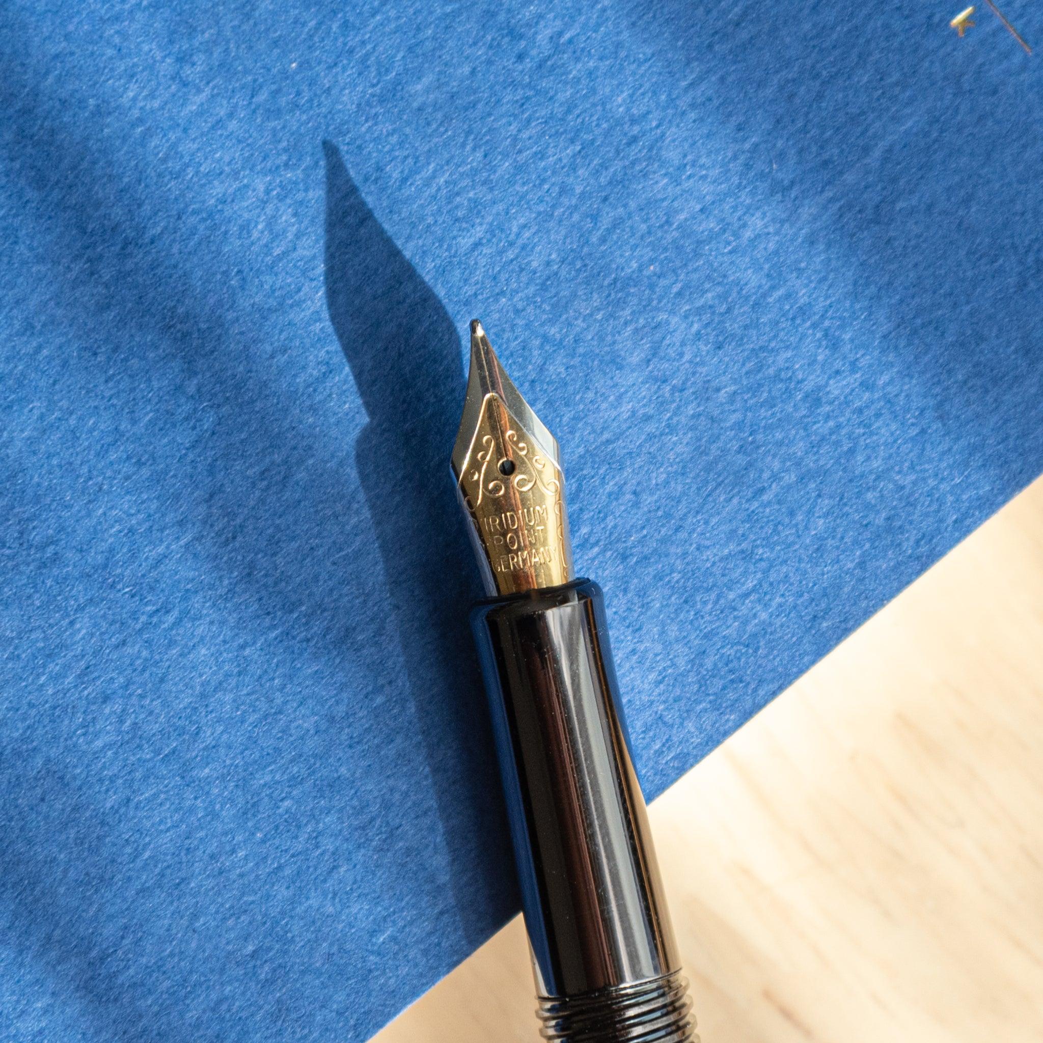 Gold nib of Japanese fountain pen with detailed engravings, resting on a vibrant blue notebook under sunlight. (91 characters)