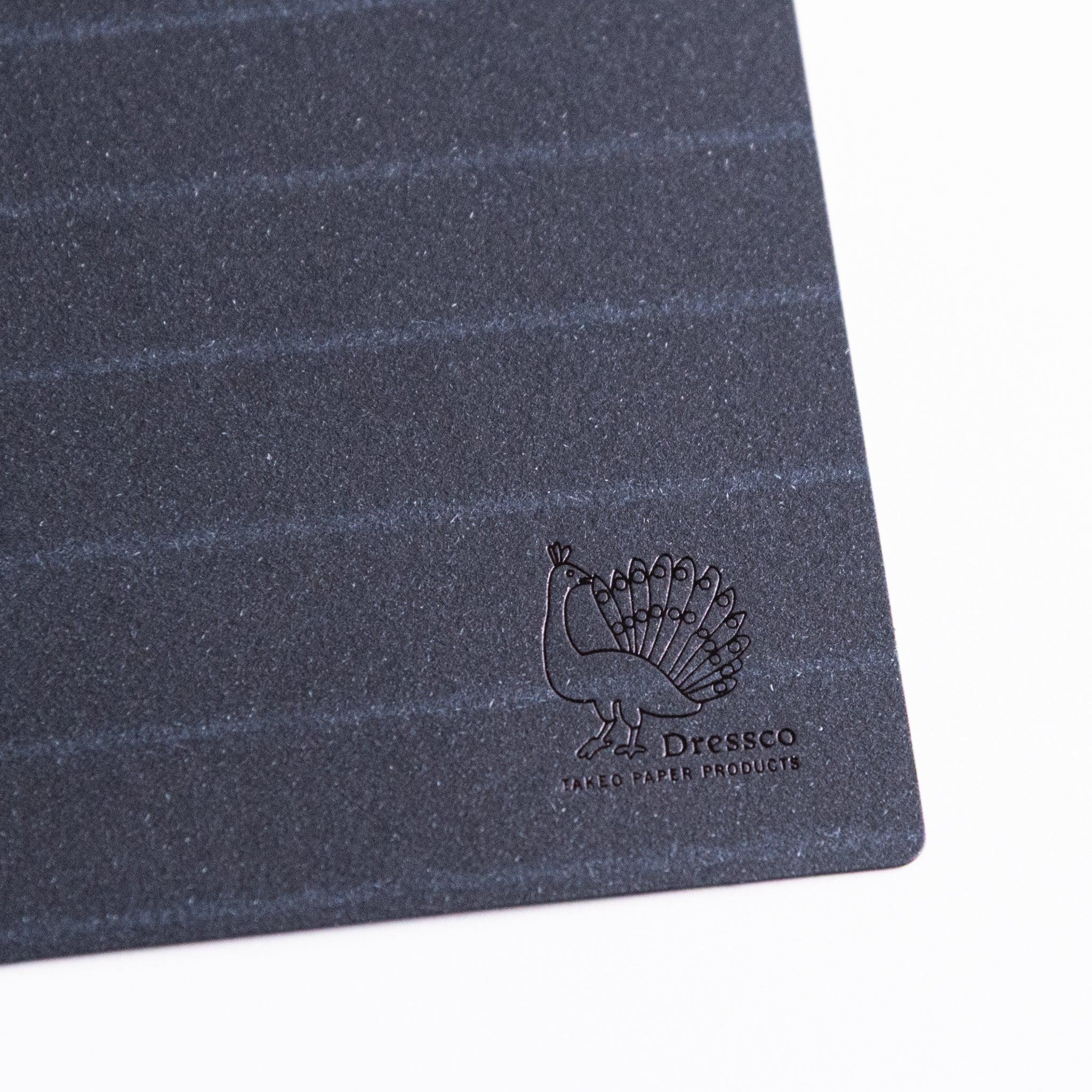 Navy paper notebook band with subtle suit pattern and Dressco peacock logo; functions as closure, bookmark, writing board.