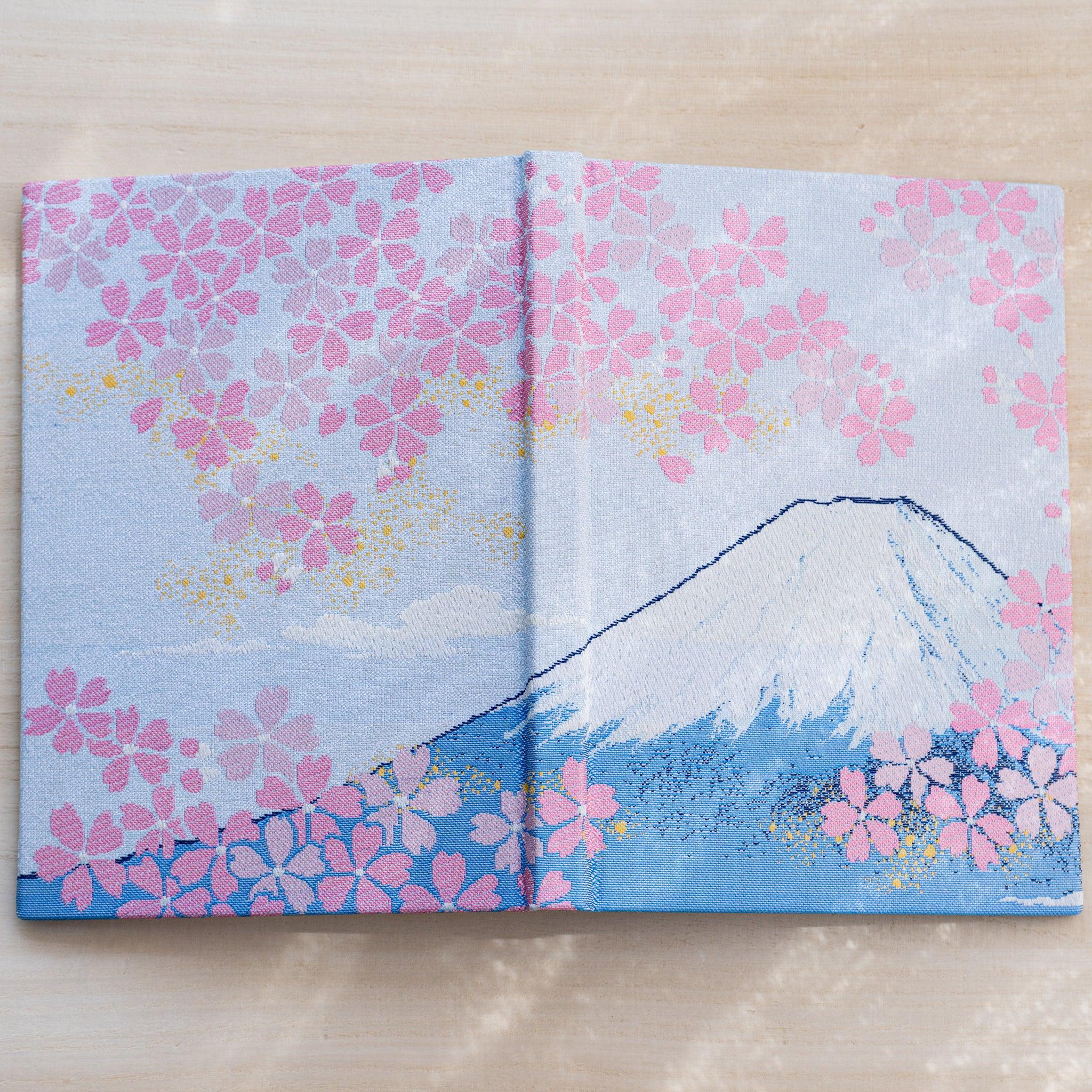 Notebook cover opened flat, woven textile spread of Mount Fuji and pink cherry blossoms on pale blue, spine at center