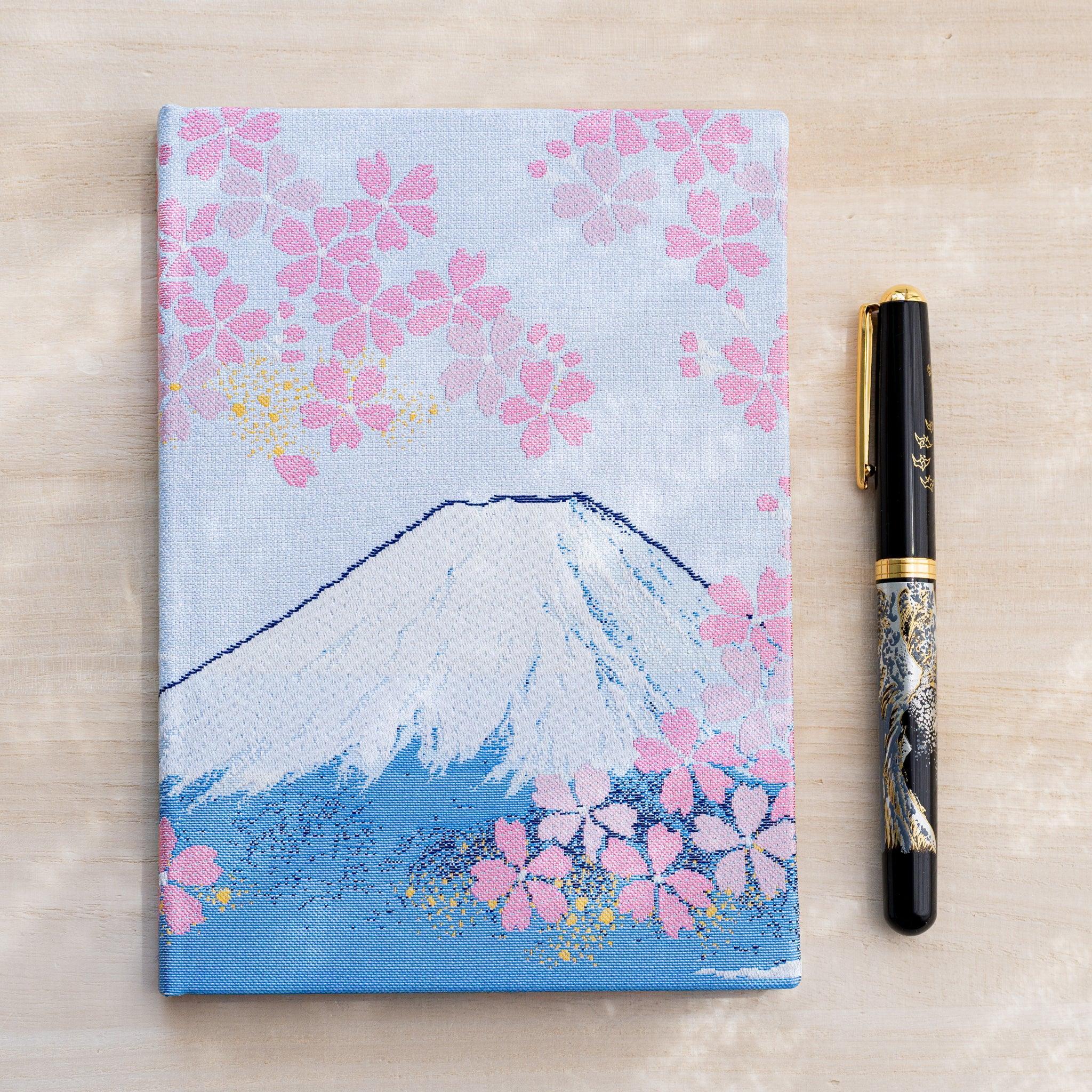 Notebook front cover with Mount Fuji and pink cherry blossoms in woven textile, black fountain pen beside it on wood