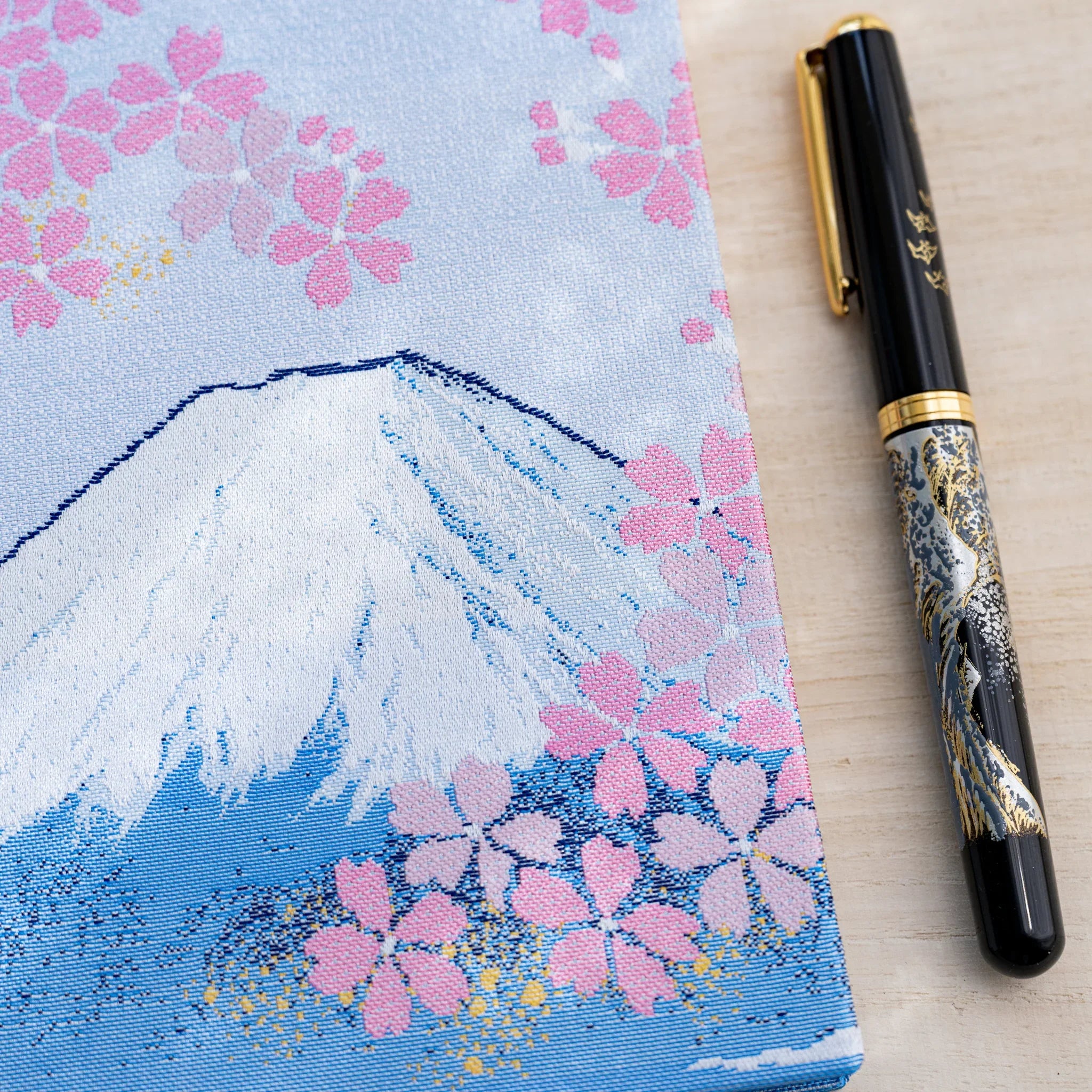 Close-up of woven textile cover showing white-capped Mount Fuji and pink cherry blossoms, black fountain pen at right