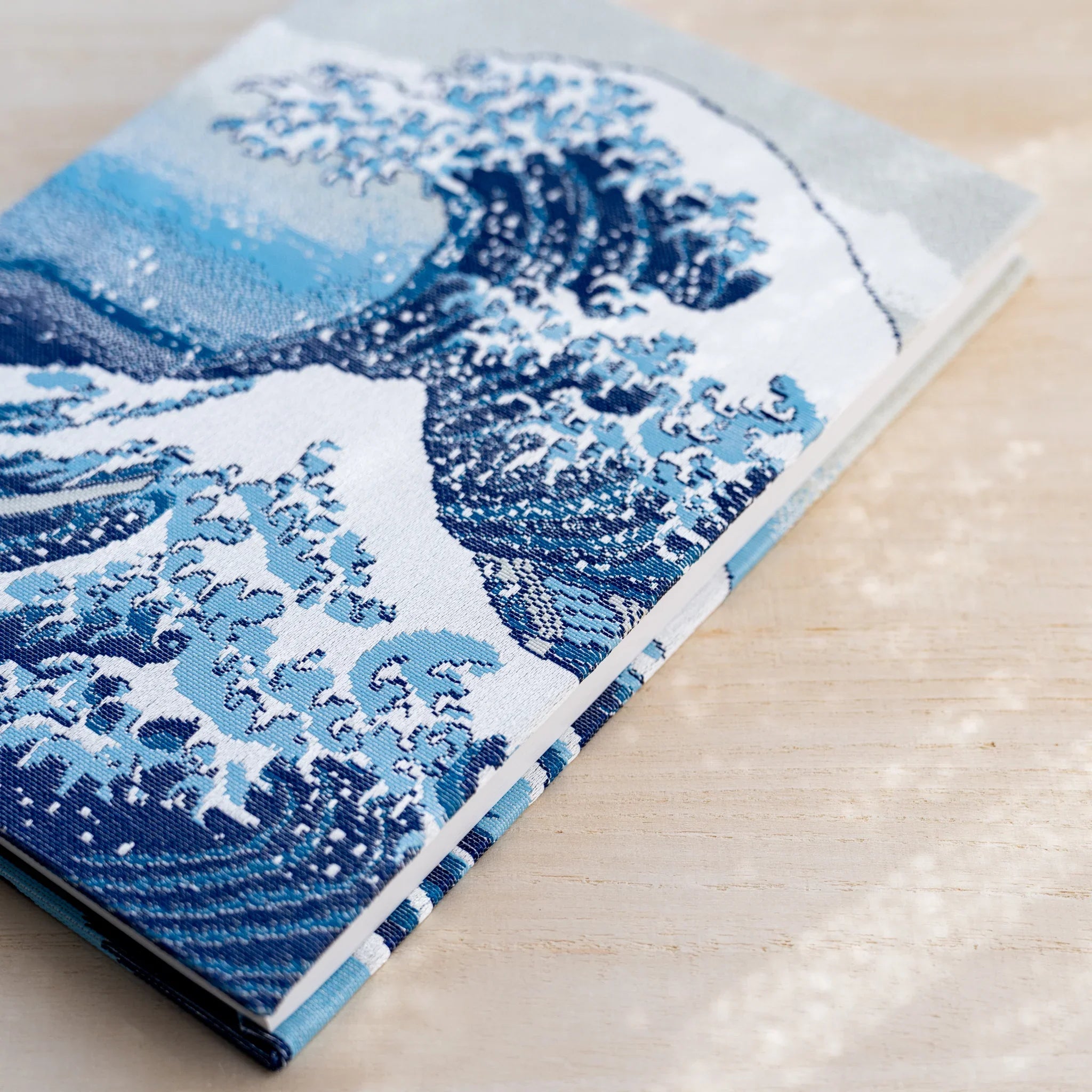 Angled view of notebook front cover, the Great Wave in blue and white woven textile on pale ground