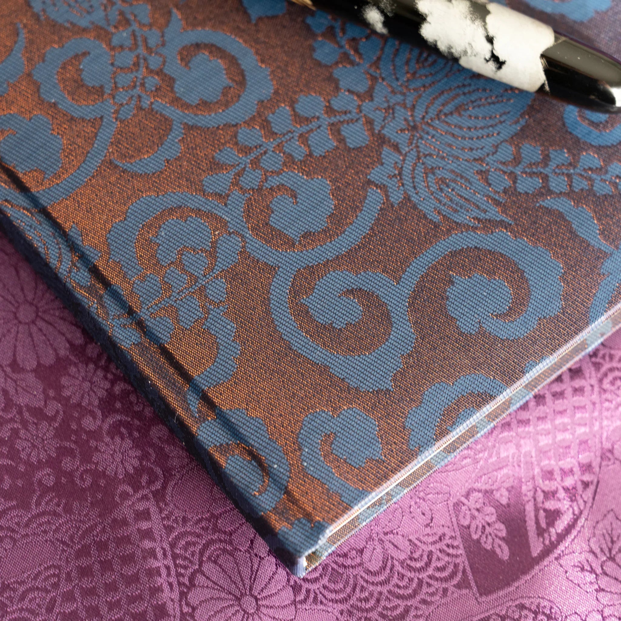 Angled corner close-up of Oni Kiri Nishijin silk cover showing blue scroll weave pattern and spine edge with fountain pen