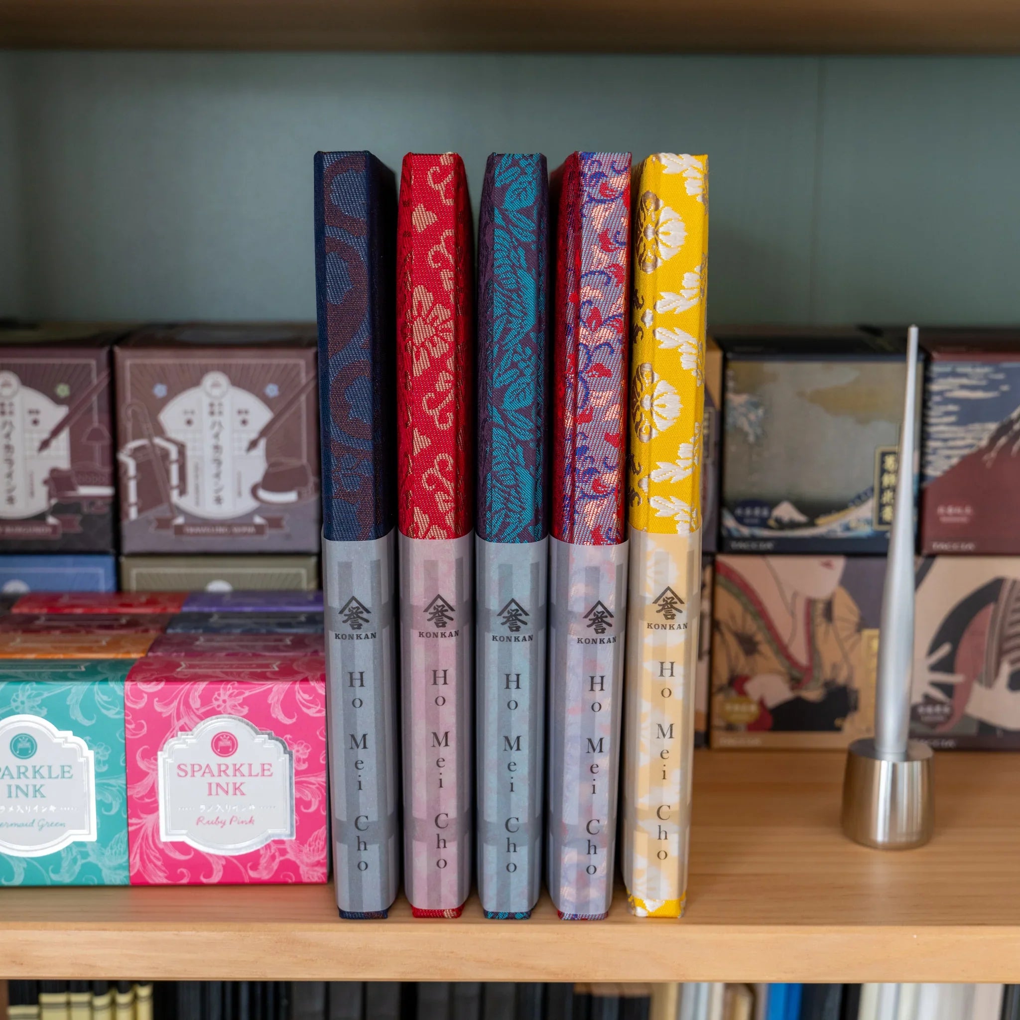 Five KONKAN Homeicho notebooks in various Nishijin silk colorways standing on wooden shelf with Japanese stationery products
