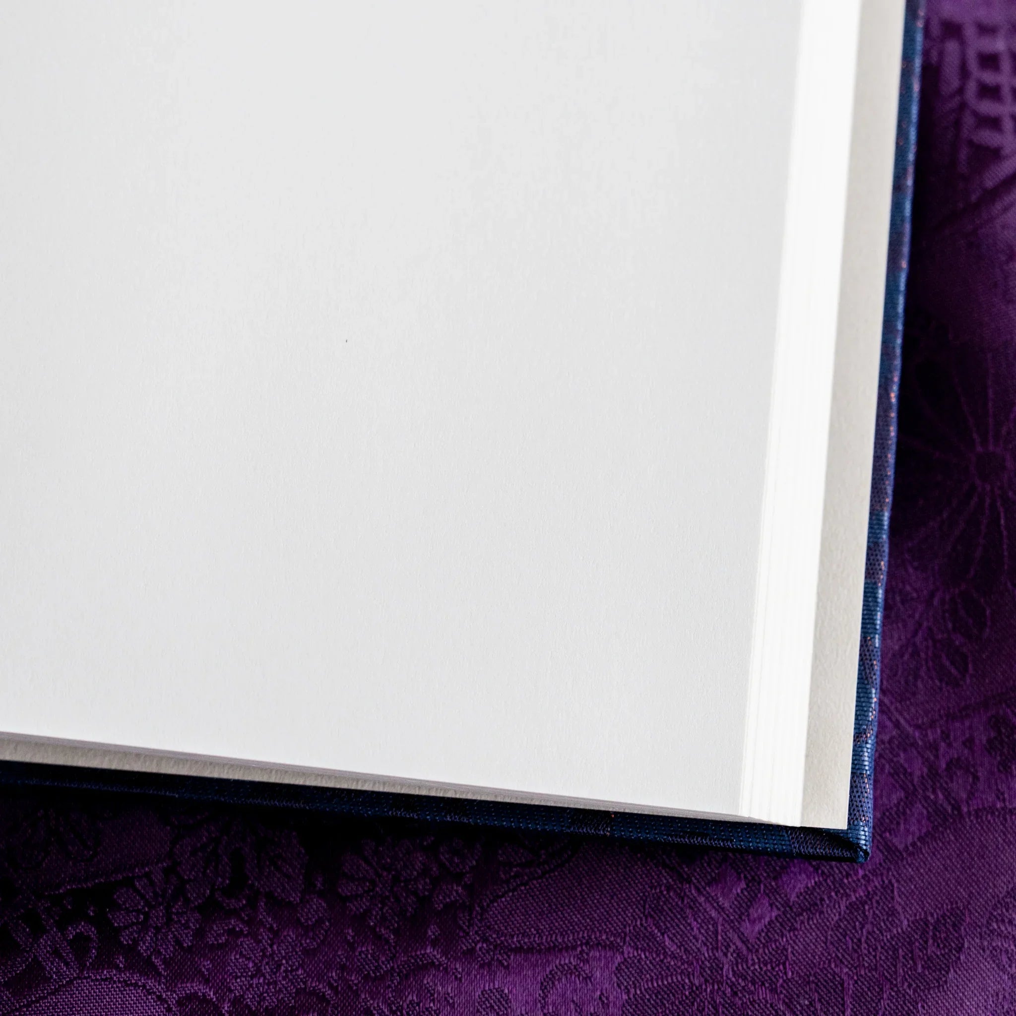 Corner close-up of Oni Kiri Nishijin Homeicho notebook showing white page stack and dark navy silk cover corner on purple