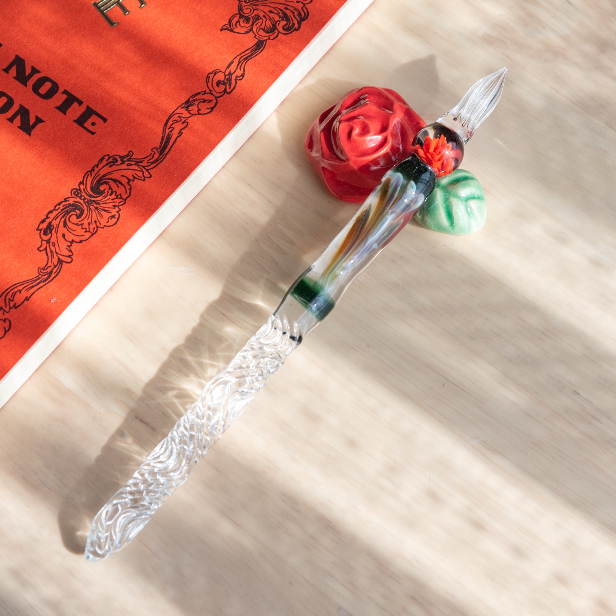 A Japanese glass dip pen with a twisted clear handle features an orange rose motif inside. It rests on a rose-shaped ceramic holder beside an orange notebook. 