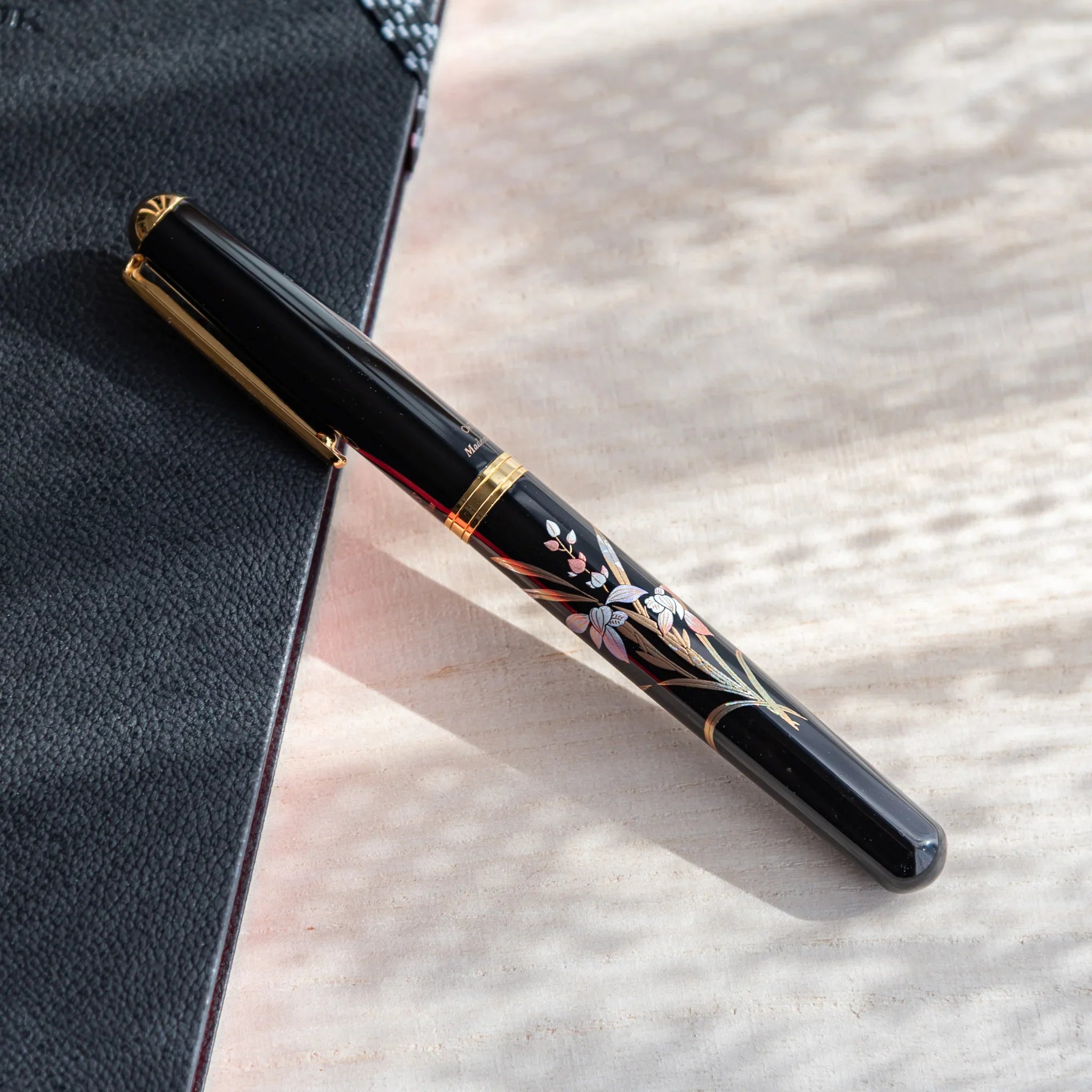 Japanese Fountain Pen with black lacquer body and orchid design, placed on a light surface beside textured dark fabric.