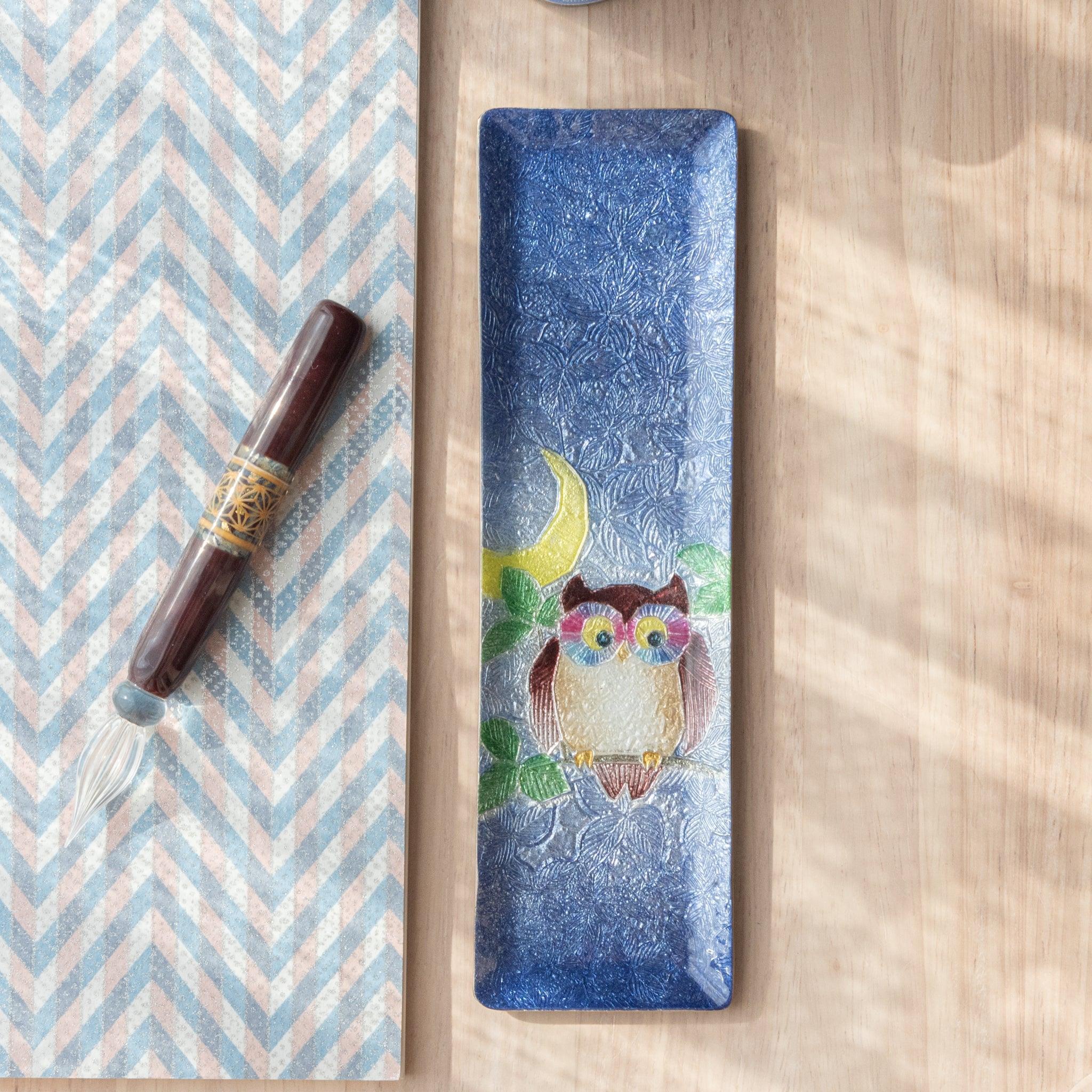 A slim blue ceramic pen tray with a colorful owl design sits beside a patterned paper and glass pen. The owl is adorned with bright colors, including a yellow crescent moon.