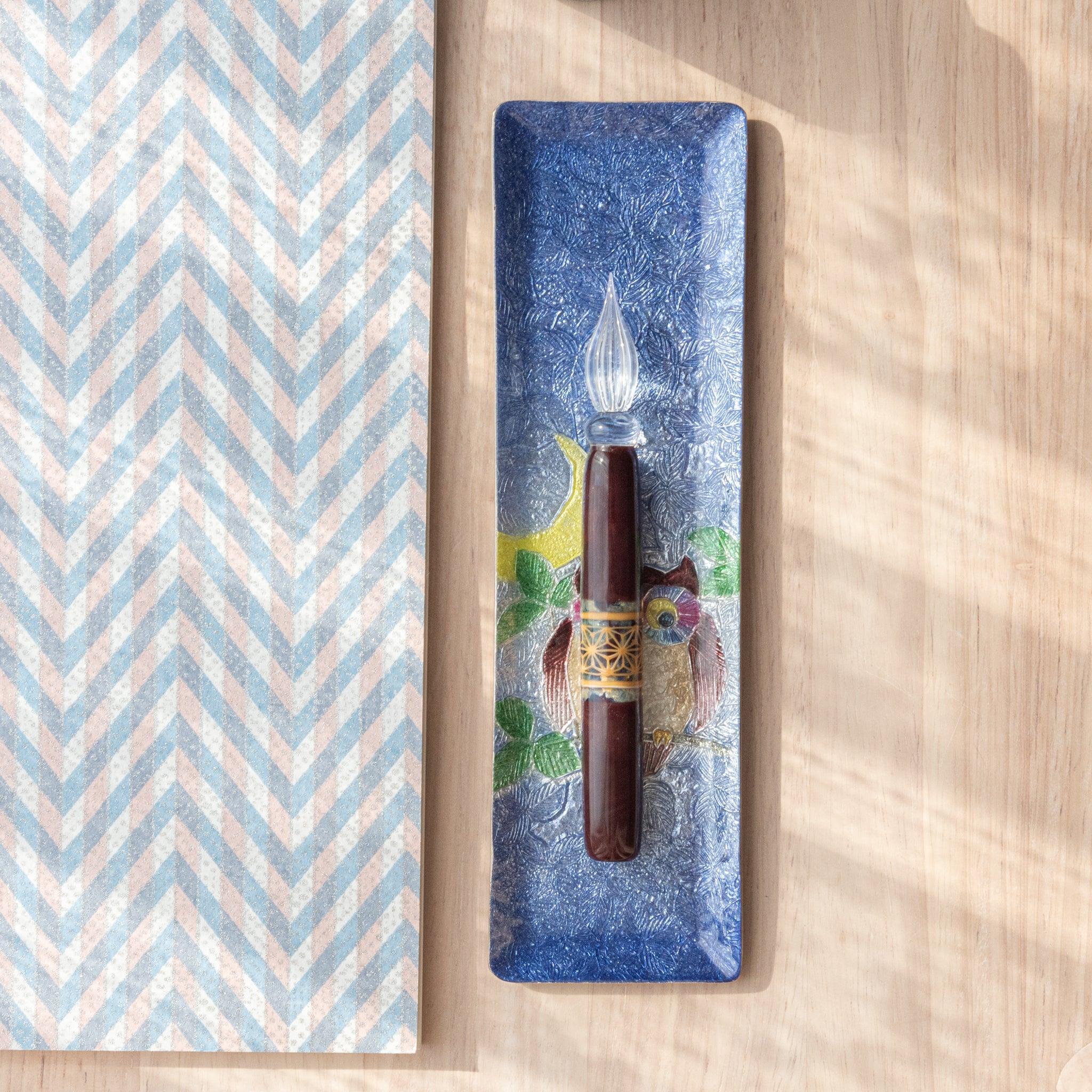 A slim blue ceramic pen tray with a colorful owl design holds a glass dip pen. The tray’s textured surface and owl with a crescent moon add artistic detail.