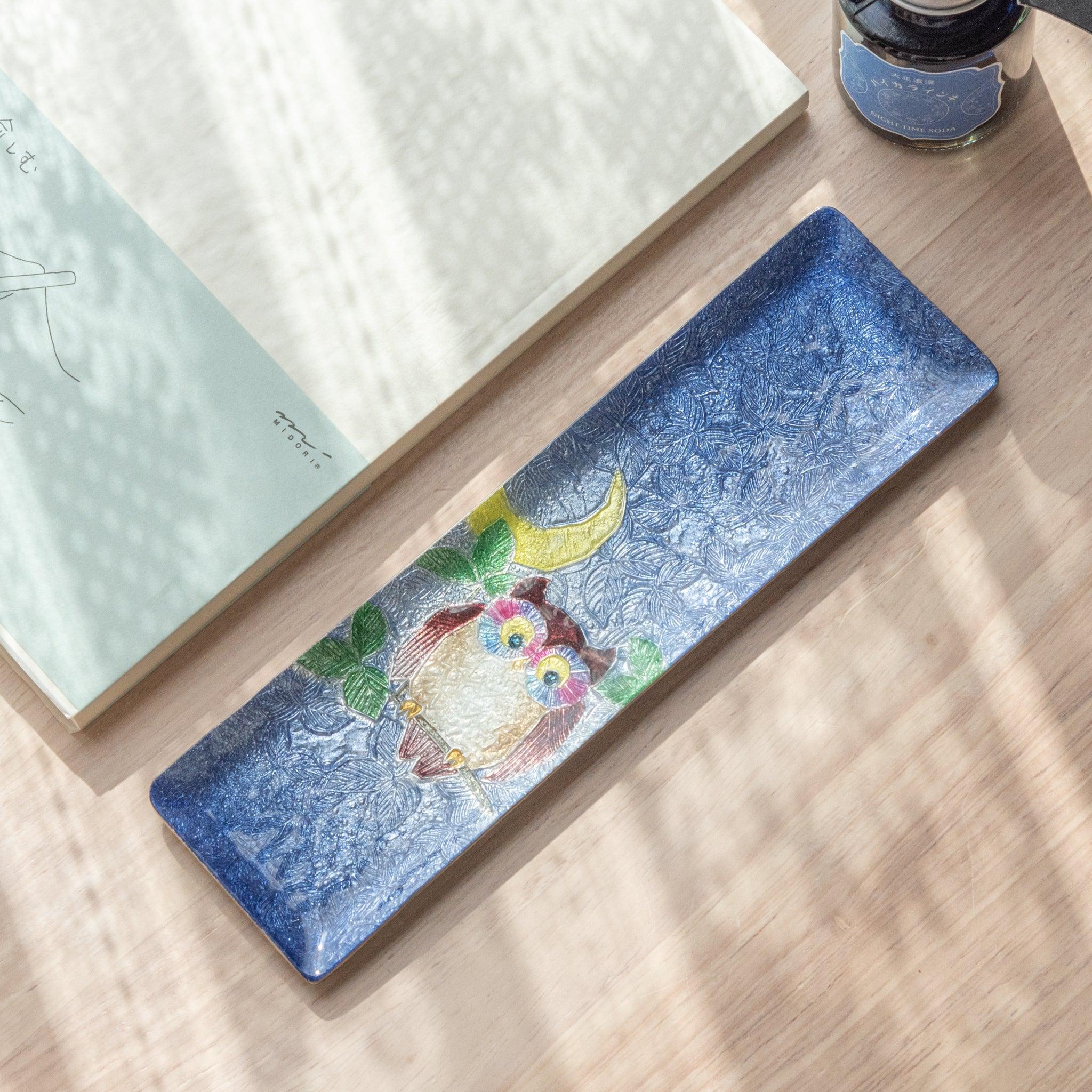 A slim blue ceramic pen tray featuring a colorful owl with a crescent moon design, placed beside an open notebook and ink bottle, on a sunlit wooden surface.