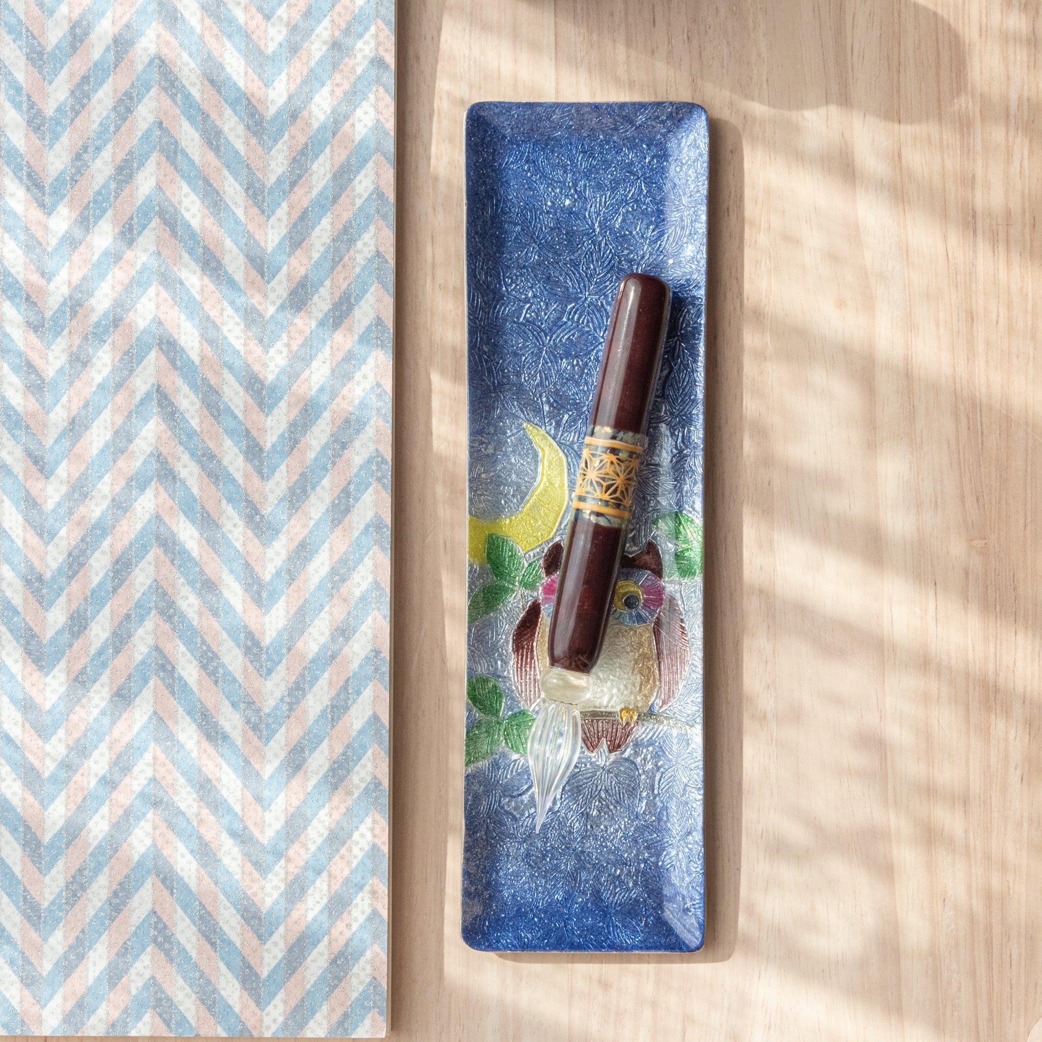 A slim blue ceramic pen tray with a colorful owl and crescent moon design holds a glass dip pen, placed beside chevron-patterned paper on a wooden surface.