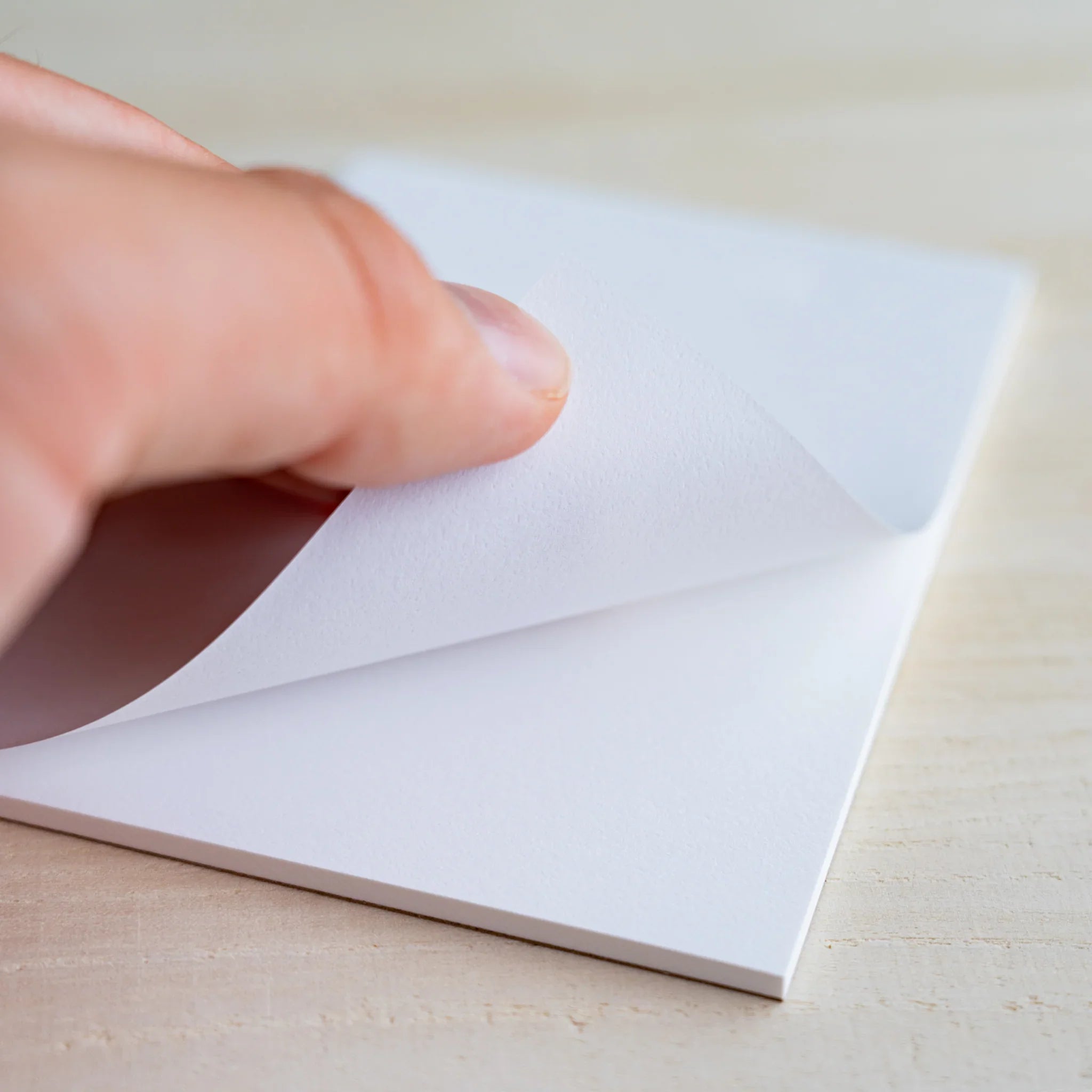 Hand lifting a white sheet from the pad showing slightly textured paper surface, on wood