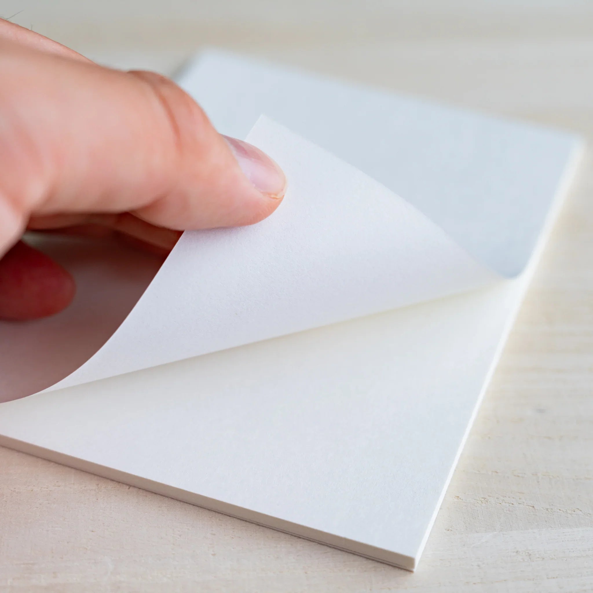 Fingers peeling back a white sheet from the bound pad to show paper texture, on wood