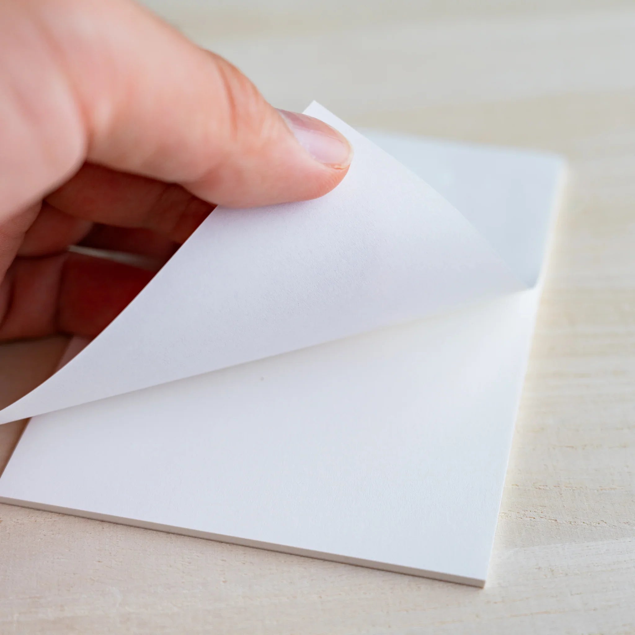 Hand lifting a white sheet from the writing pad to show paper thickness, on light wood