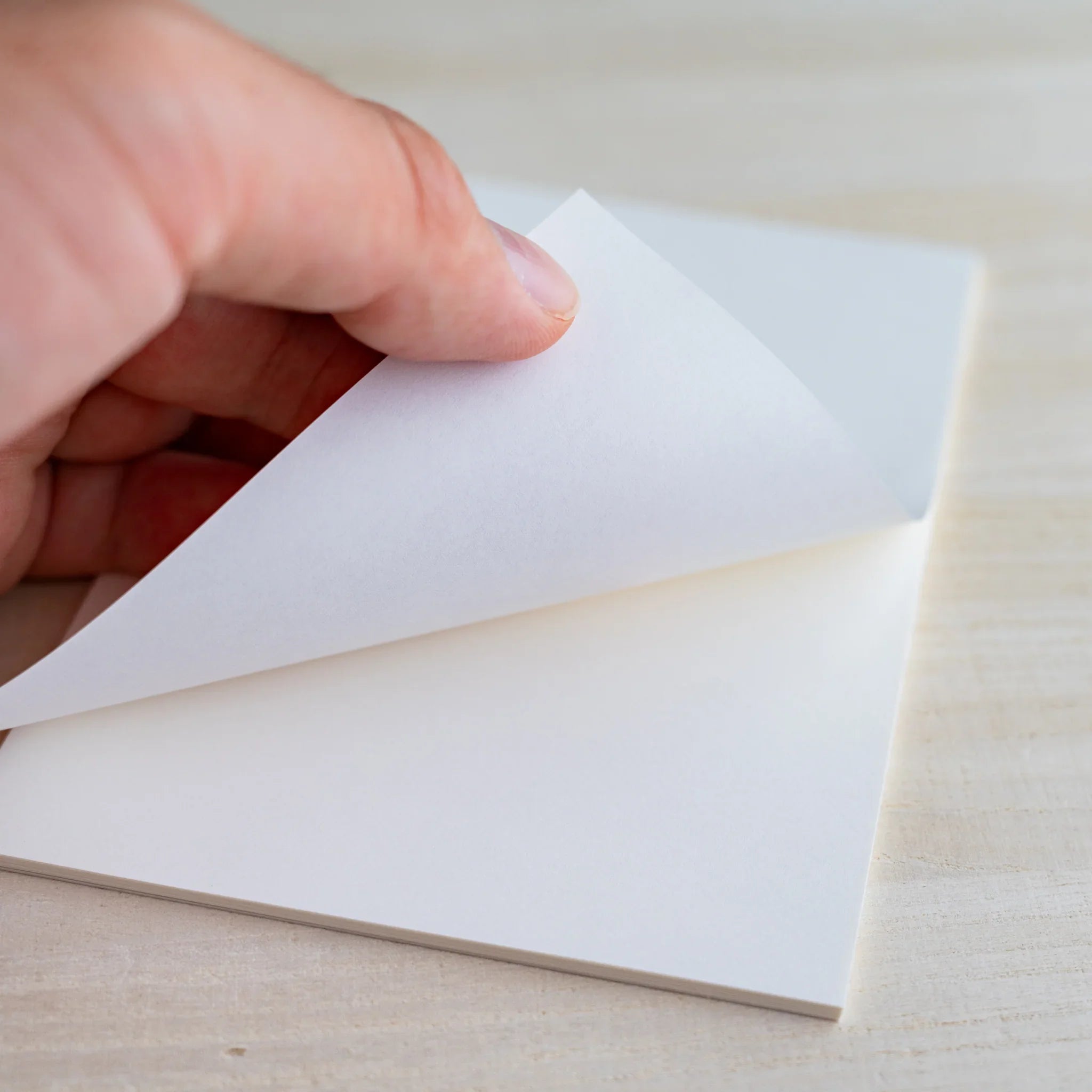 Hand peeling back a white sheet from the bound writing pad, light wood background