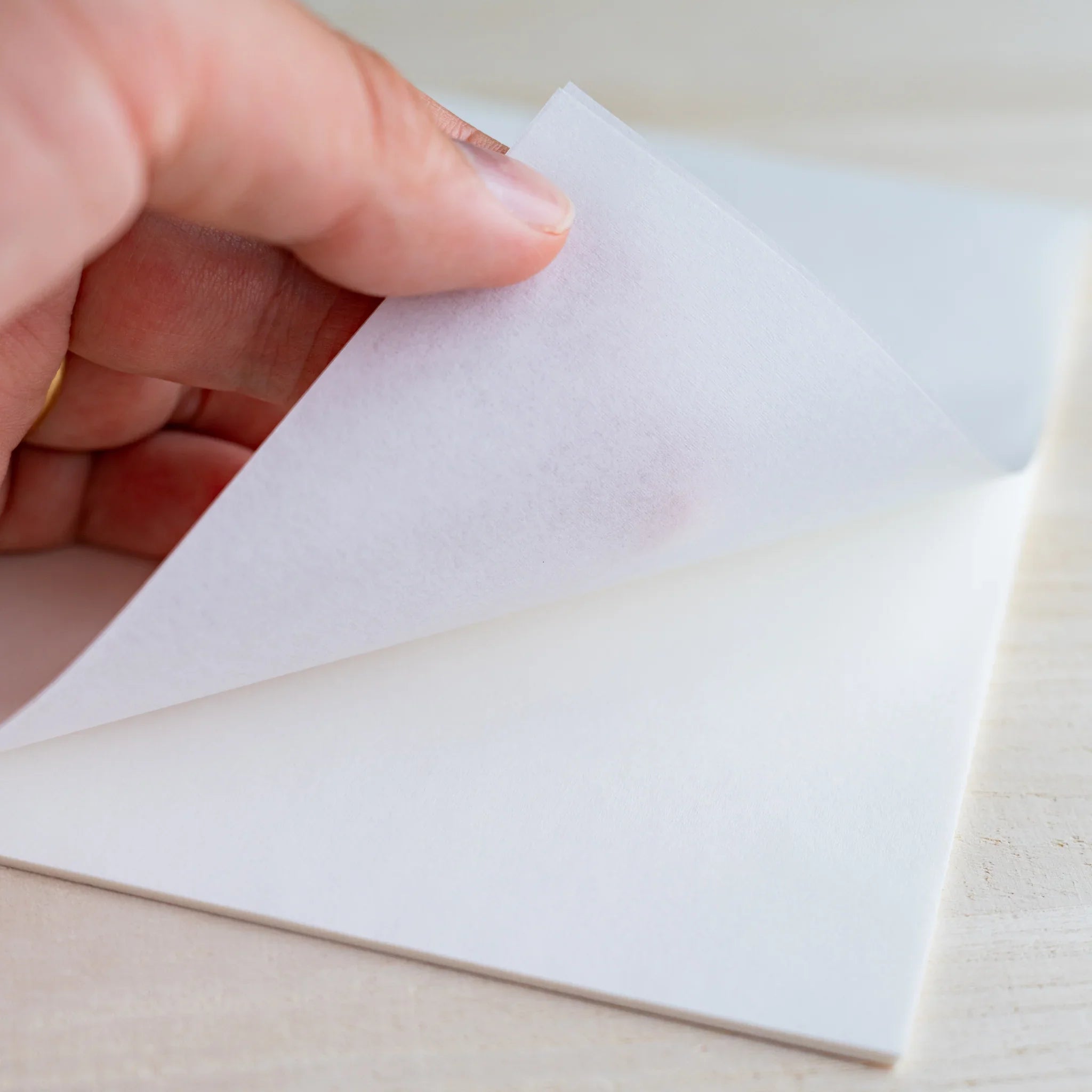 Hand lifting a thin onionskin sheet, light passing through the translucent paper, on wood