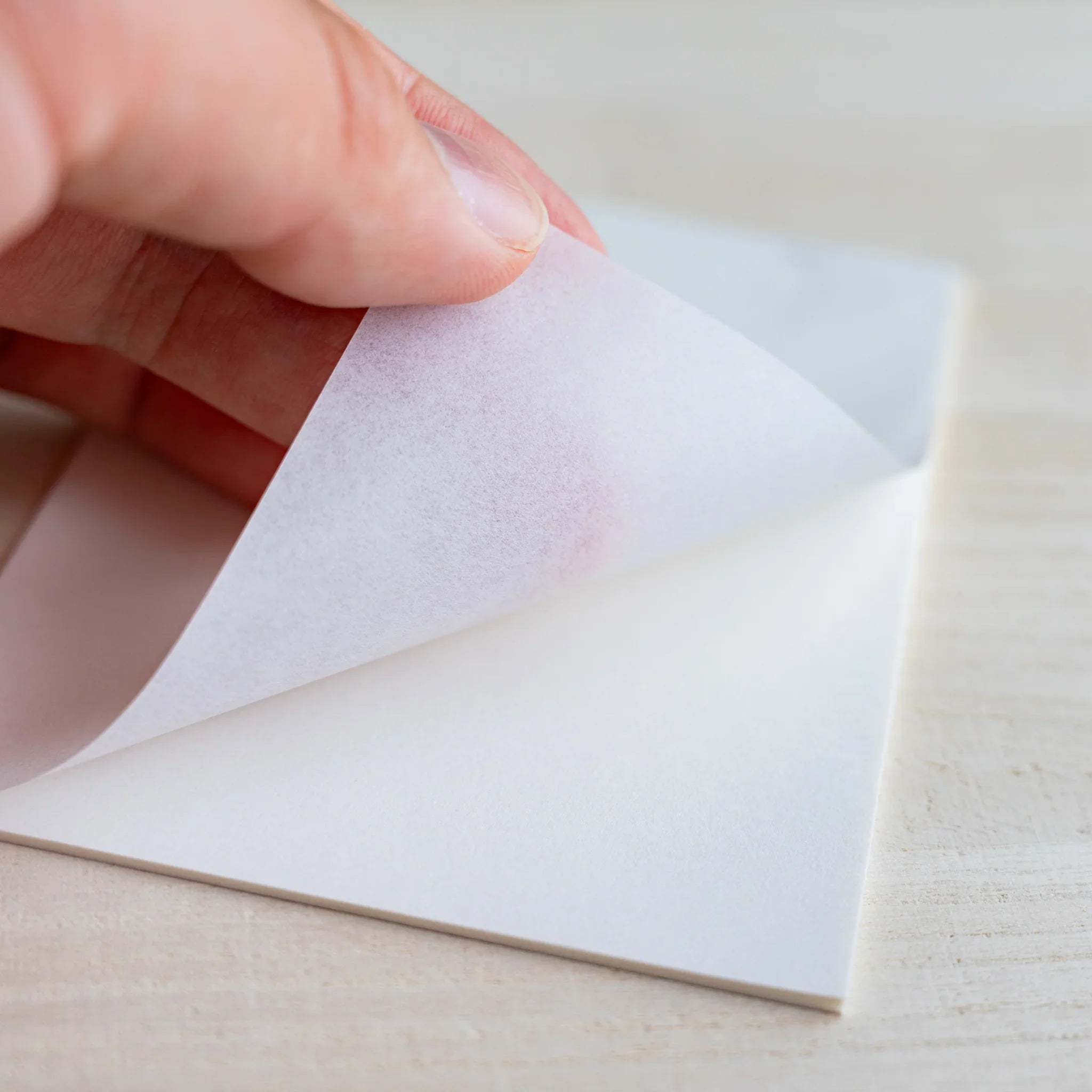 Hand lifting a thin onionskin sheet, fingers faintly visible through the translucent paper, on wood