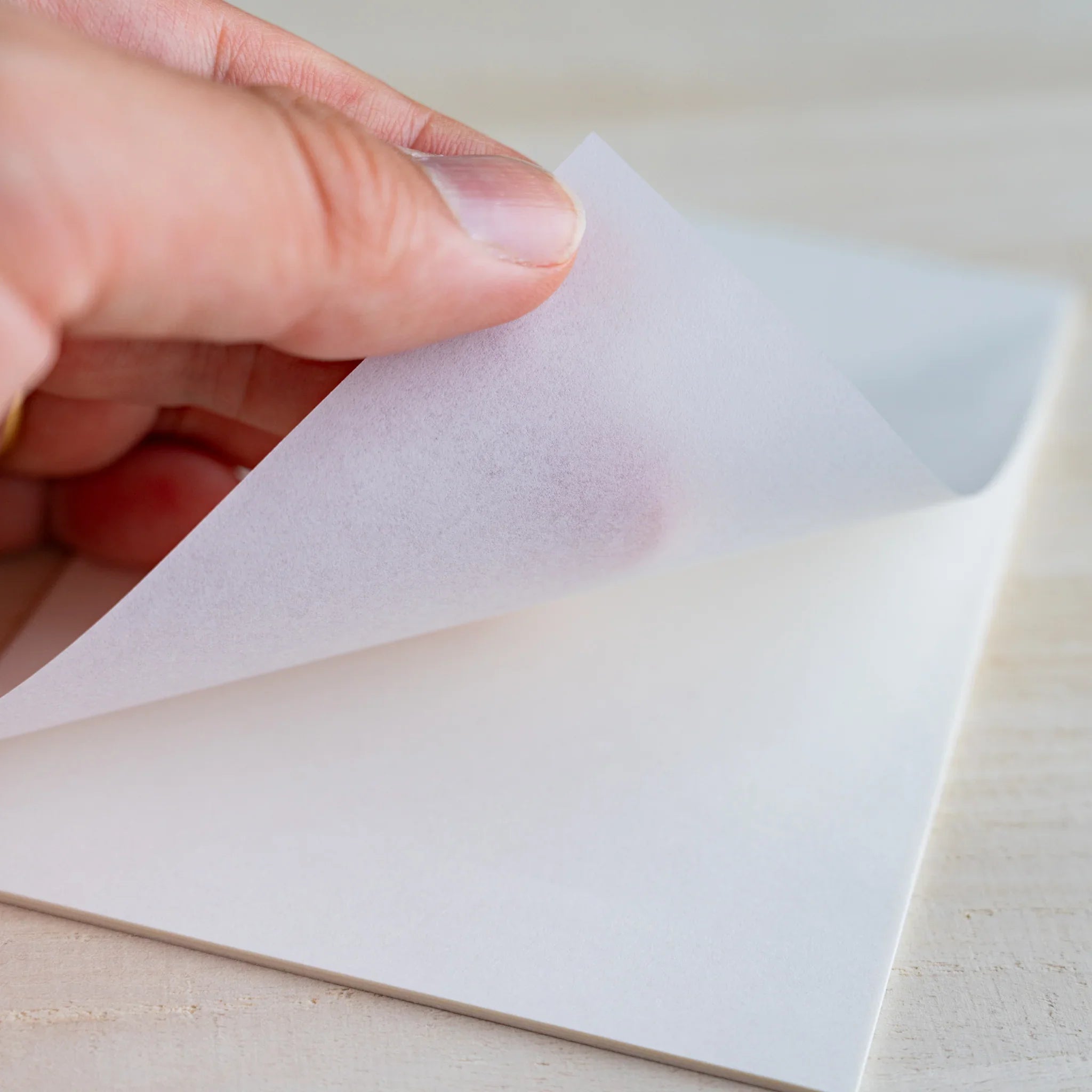 Fingers peeling a thin onionskin sheet, showing translucency with hand visible underneath, on wood