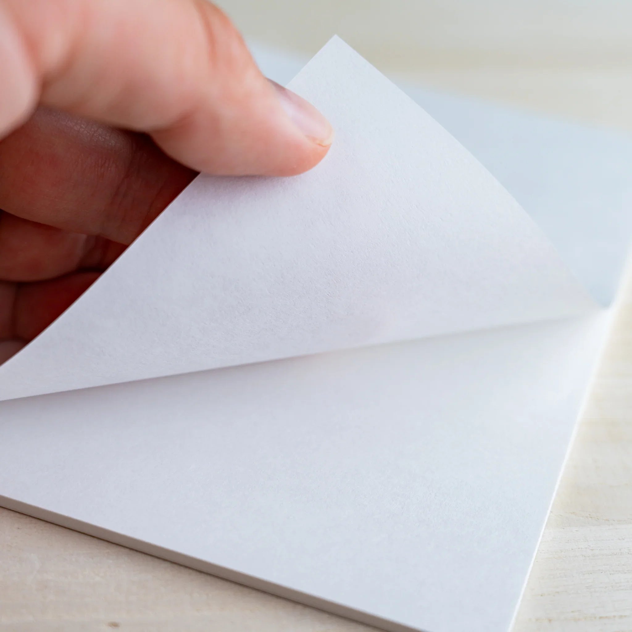 Hand lifting a white sheet from the writing pad to show single-sheet thickness, on wood