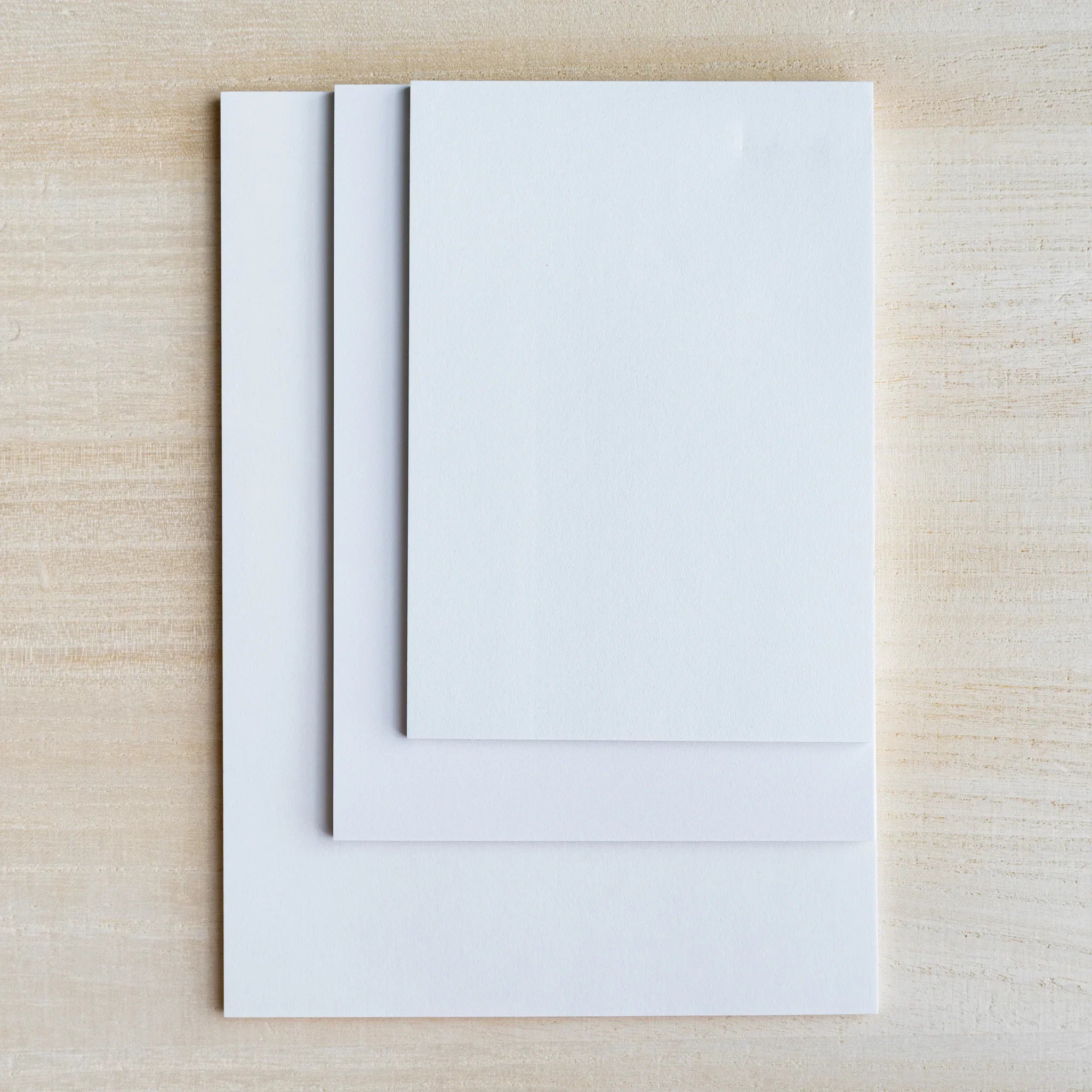 Three blank white sample sheets stacked at different sizes on light wood surface