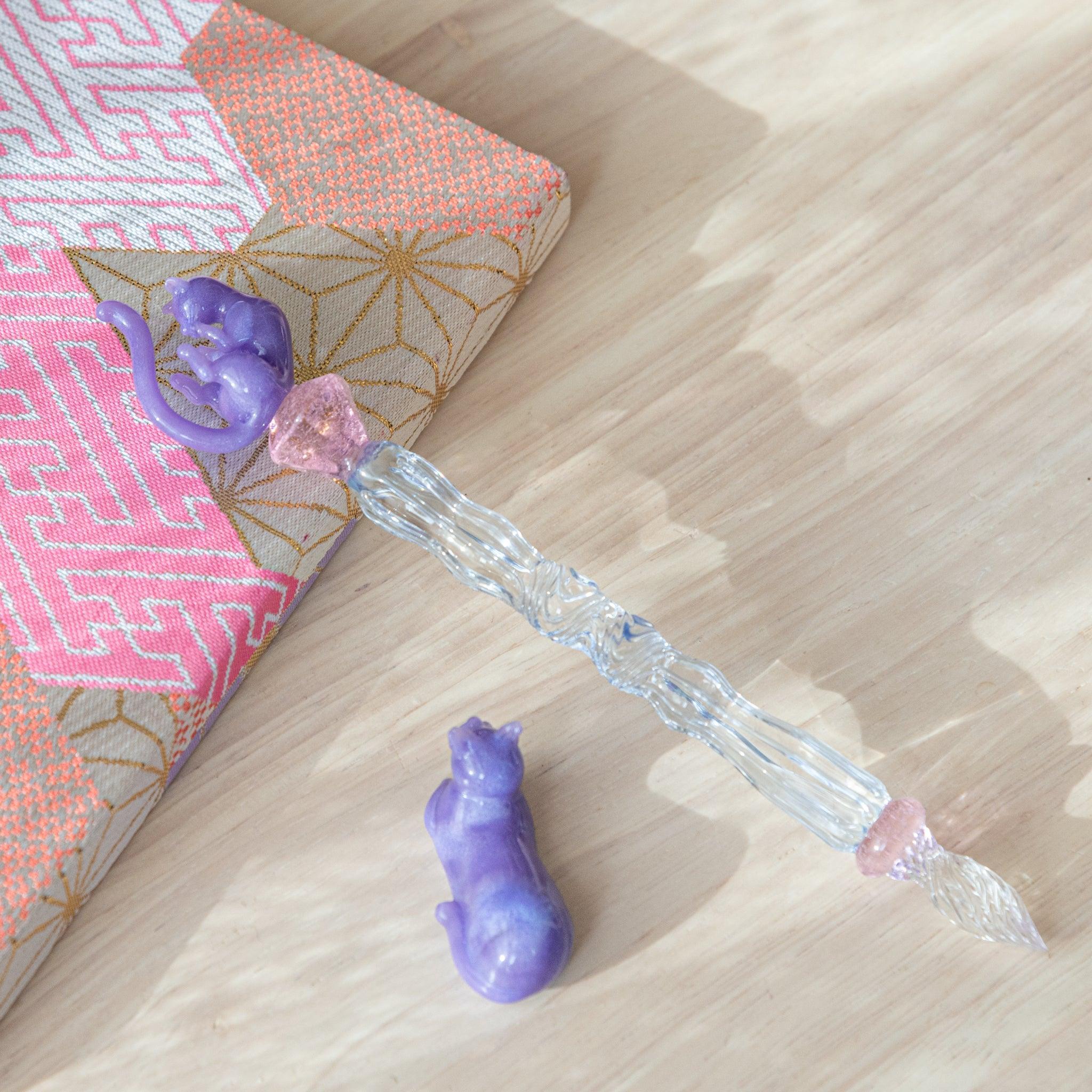 Glass pen with intricate blue shaft and purple cat topper, accompanied by a matching purple pen rest. Ideal for collectors.