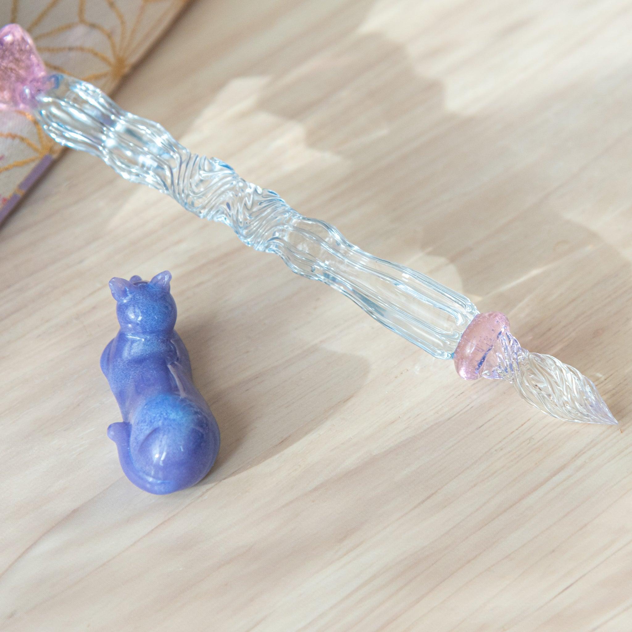 Close-up of a glass pen with a blue shaft and purple cat topper. The pink charm between the shaft and nib adds elegance.