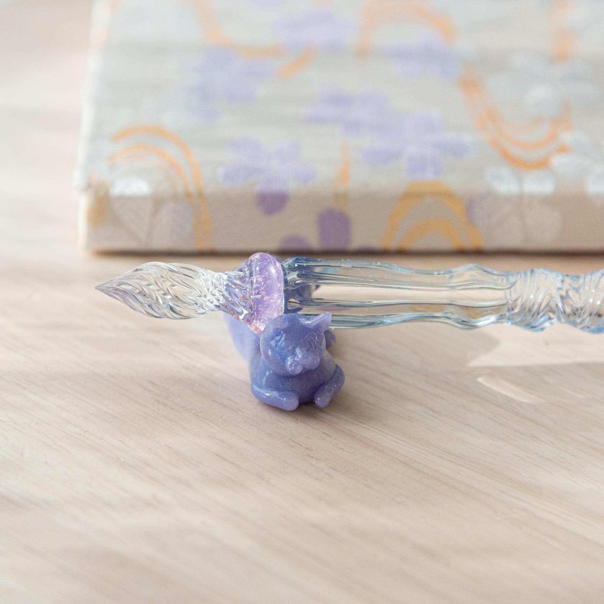 Close-up of a glass pen with a blue shaft and purple cat topper. The pink charm between the shaft and nib adds elegance.