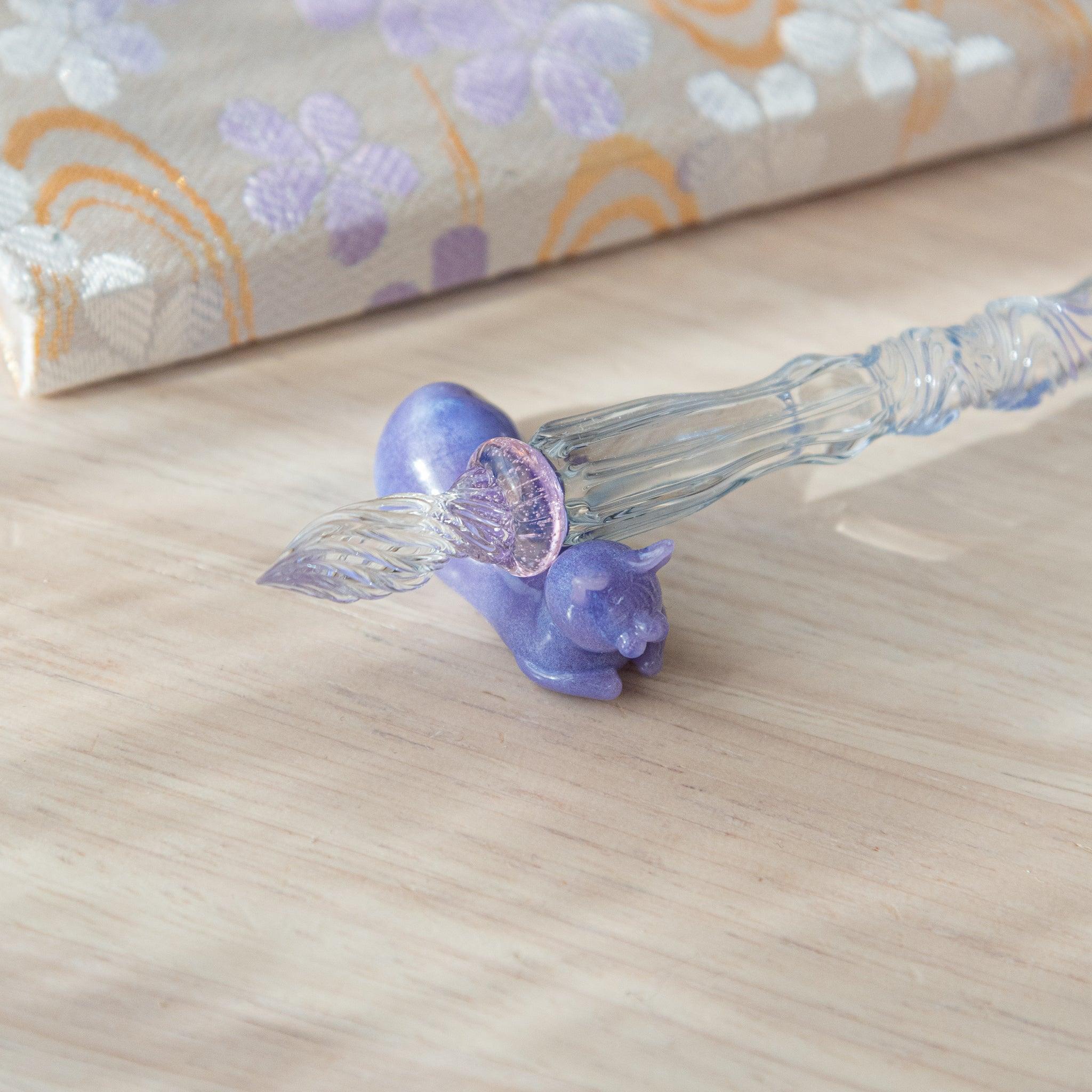 Close-up of a glass pen with a blue shaft and purple cat topper. The pink charm between the shaft and nib adds elegance.