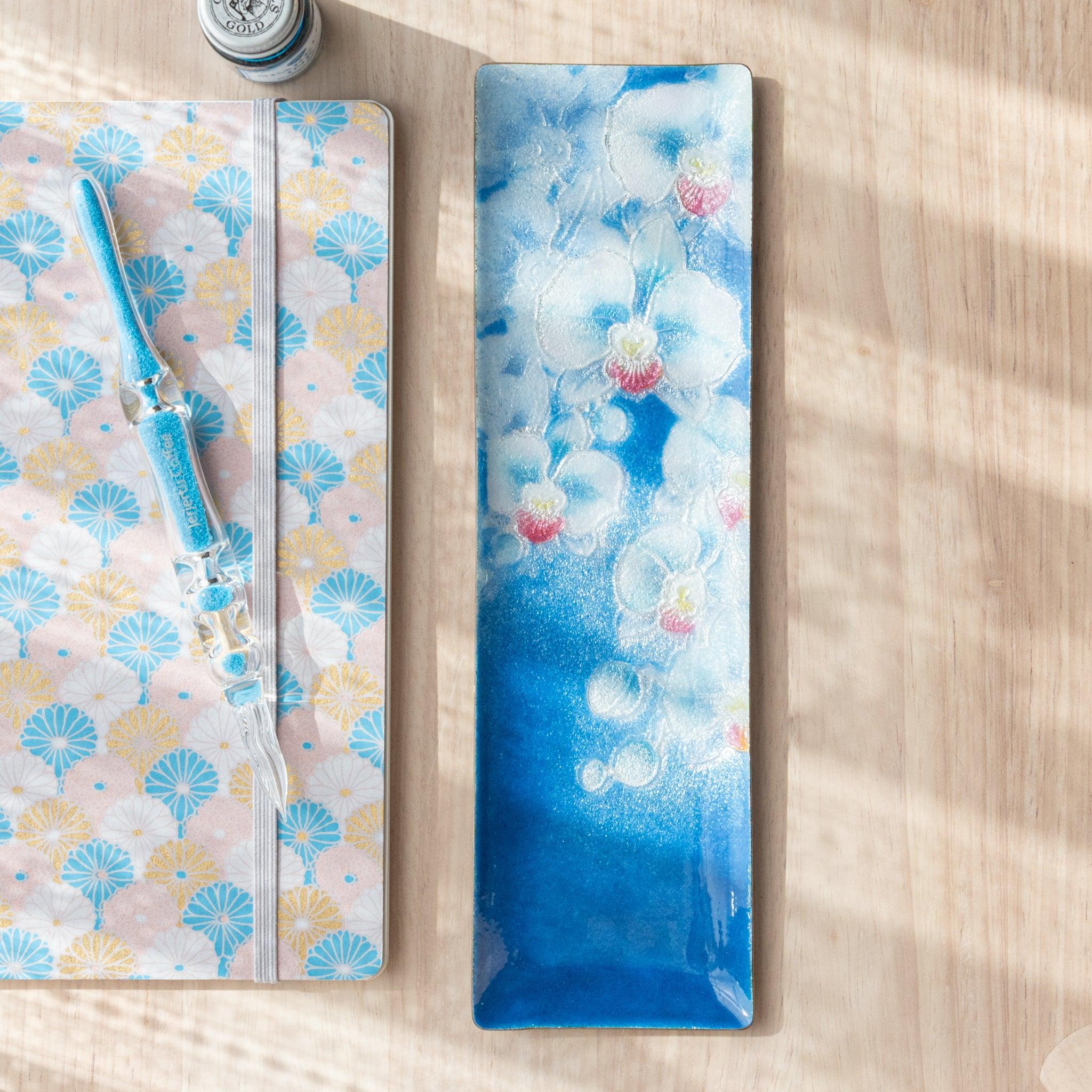 Blue rectangular tray with delicate white and pink orchid design, placed beside a pastel fan-patterned notebook and matching blue glass pen.