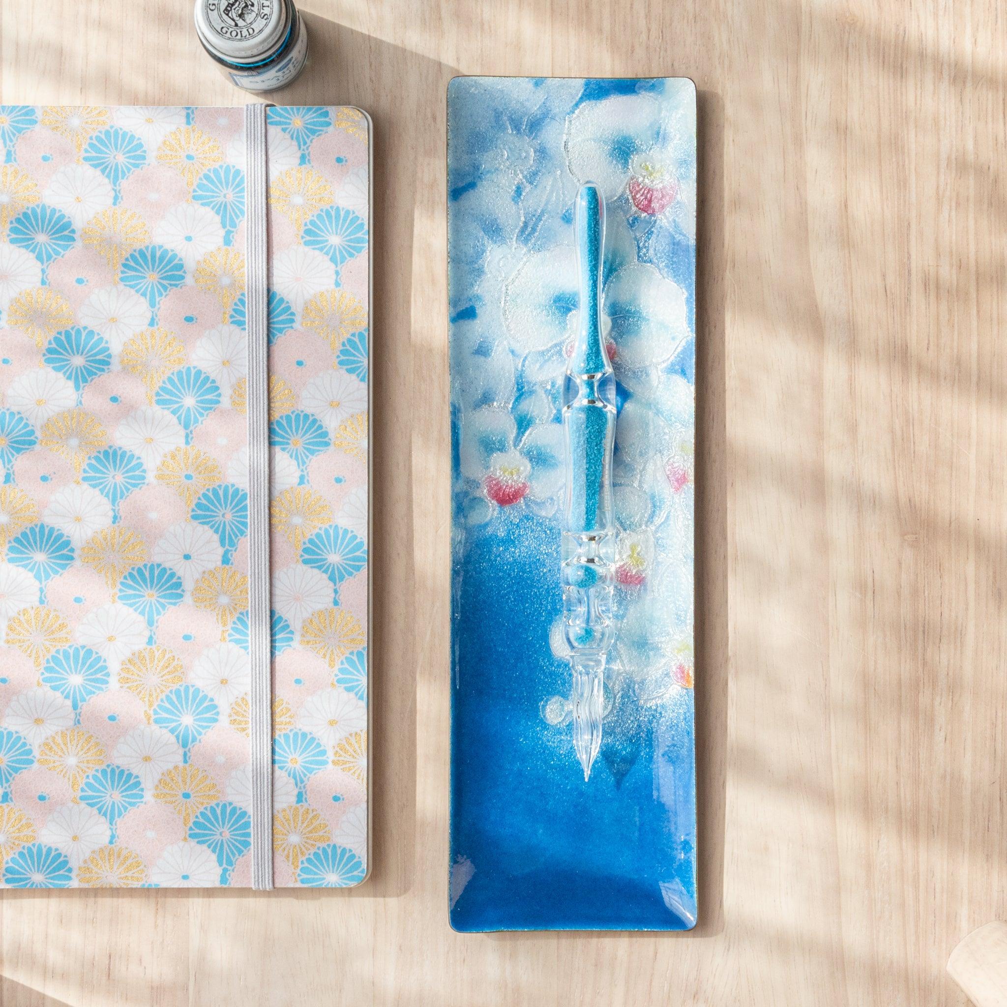 Blue rectangular tray with orchid pattern and a blue glass pen on top, alongside a fan-patterned notebook on a light wooden surface.