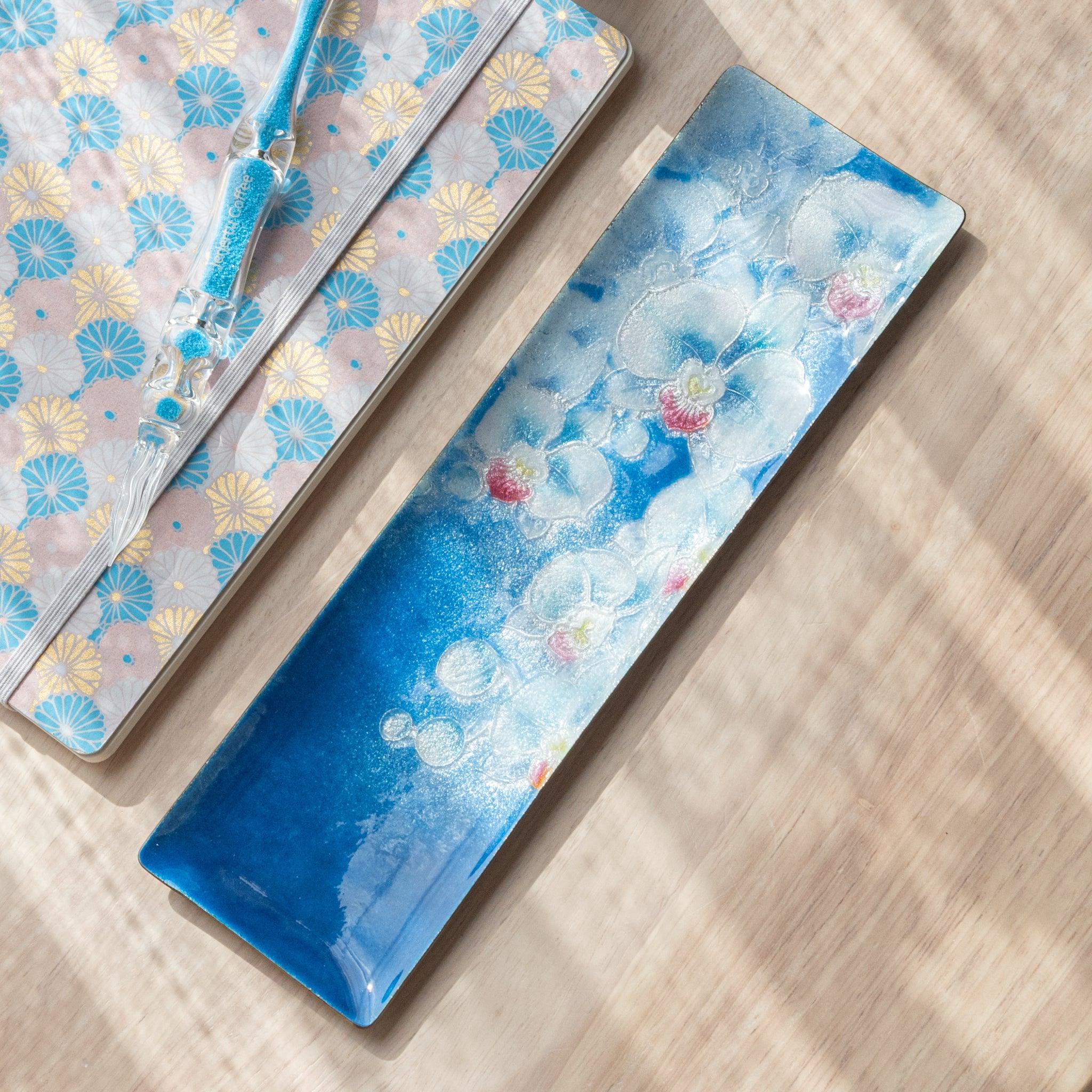 Blue rectangular tray with a white and pink orchid design, placed at an angle next to a pastel-patterned notebook and blue glass pen on a wooden surface.