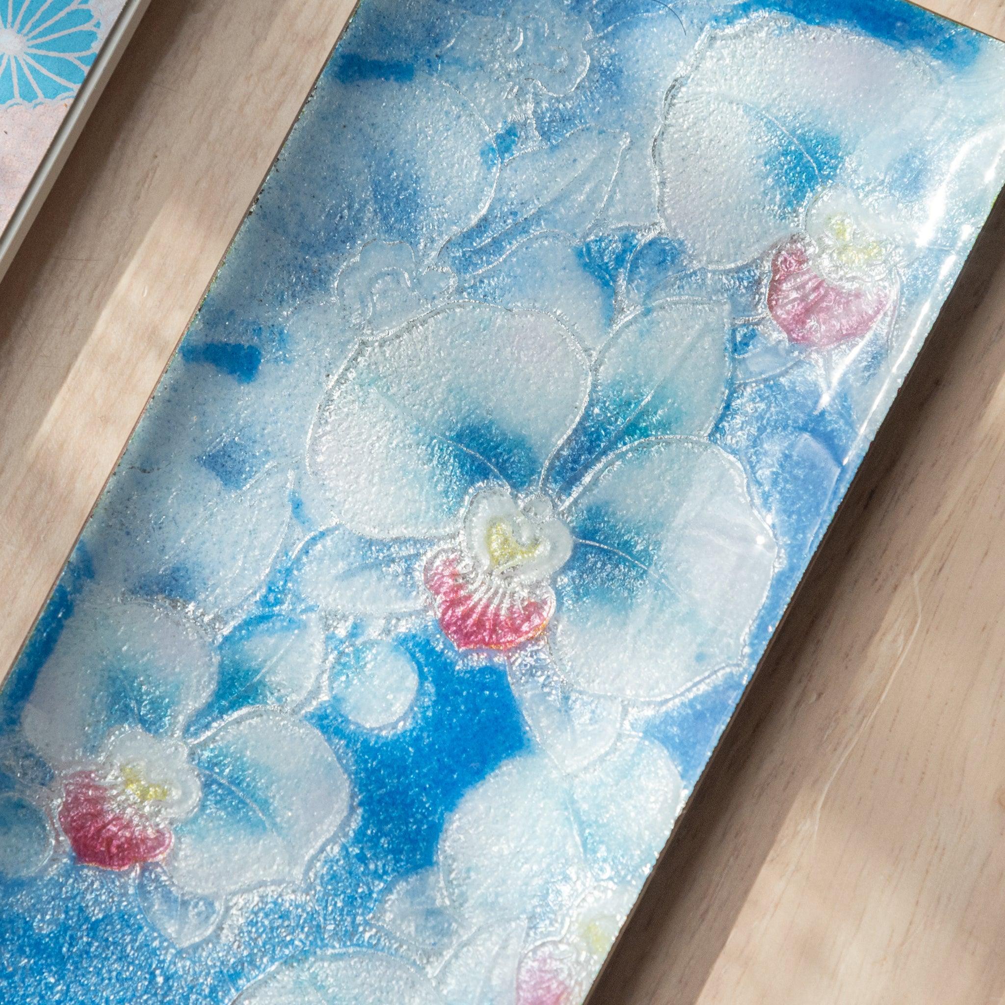 Close-up of a blue rectangular tray with white orchids and pink centers, showing fine detailing and texture on a wooden surface.