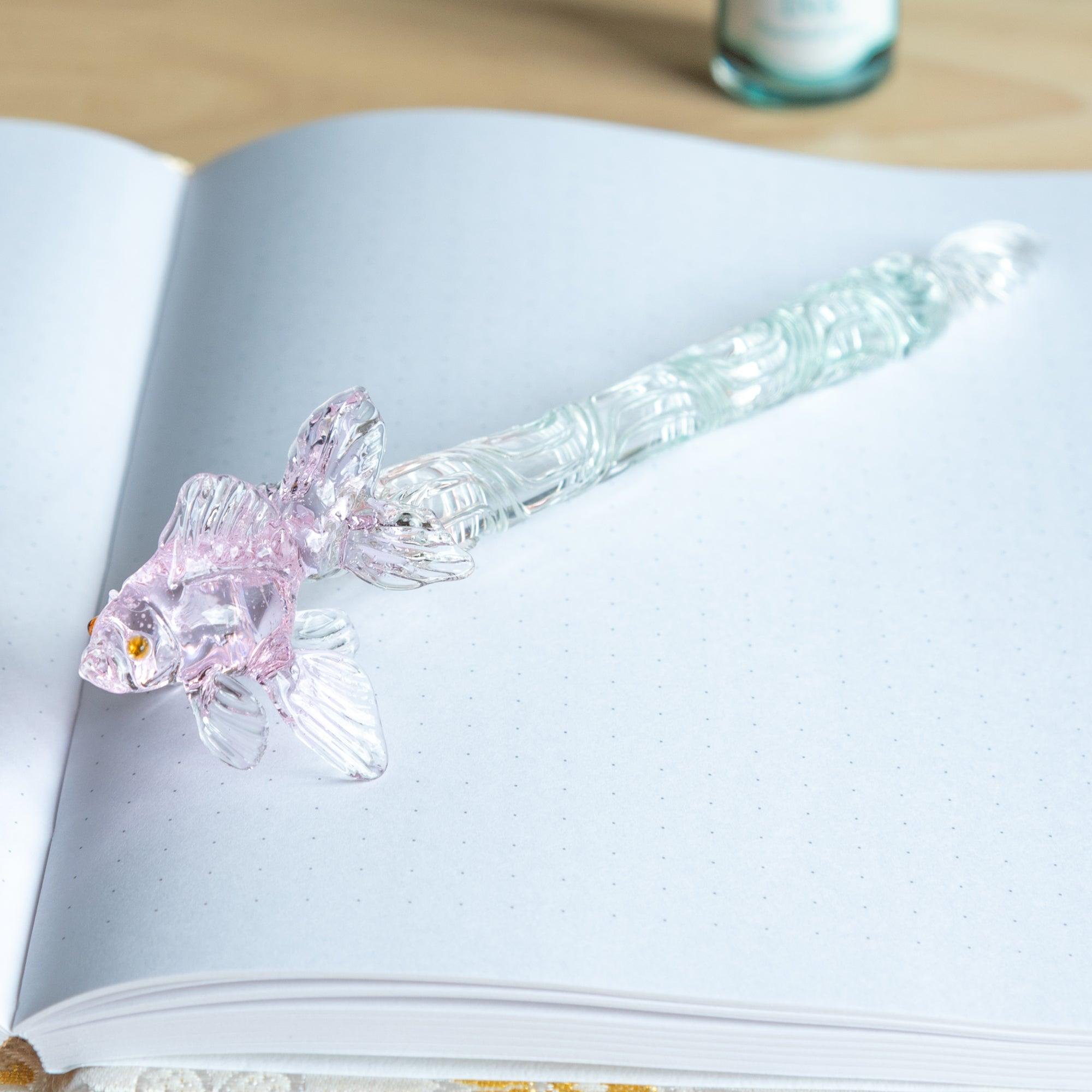 A close-up of a delicate pink goldfish-shaped glass pen, with intricate details of the fish’s fins and body, placed near a bottle of sparkling ink on a desk.