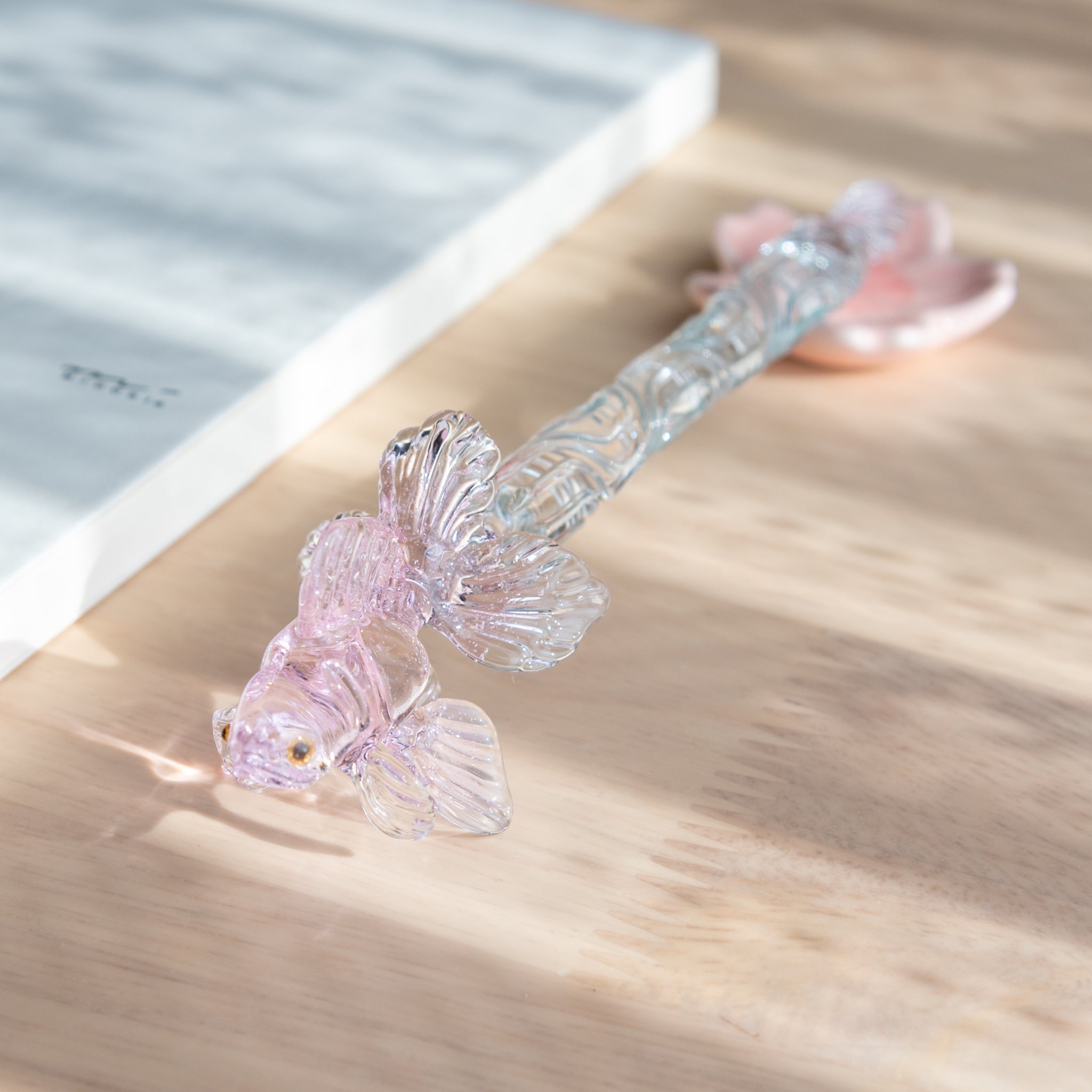 A Japanese glass dip pen with a pink goldfish motif and intricate glass details rests on a pink maple leaf-shaped holder beside an MD notebook on a wooden desk.  