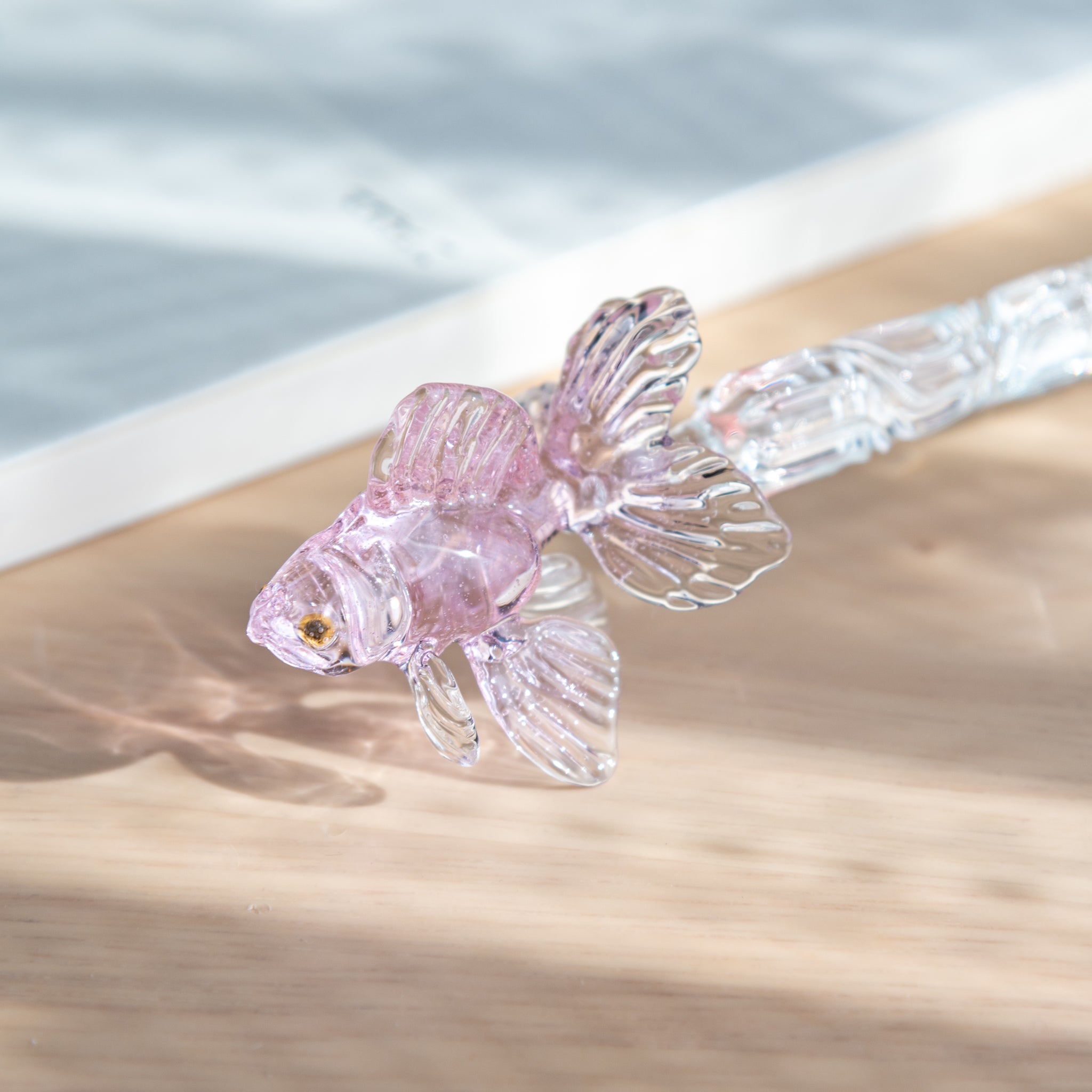 A close-up of a Japanese glass dip pen featuring a delicate pink goldfish motif with translucent fins, placed near an MD notebook on a wooden desk.