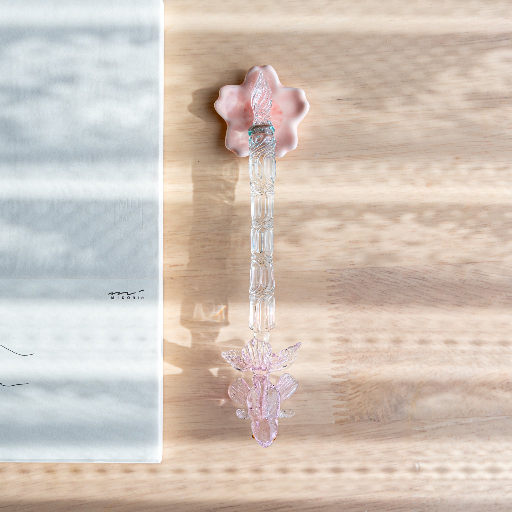 A Japanese glass dip pen with a pink goldfish motif and textured clear handle rests on a pink maple leaf-shaped holder beside an MD notebook on a wooden desk.