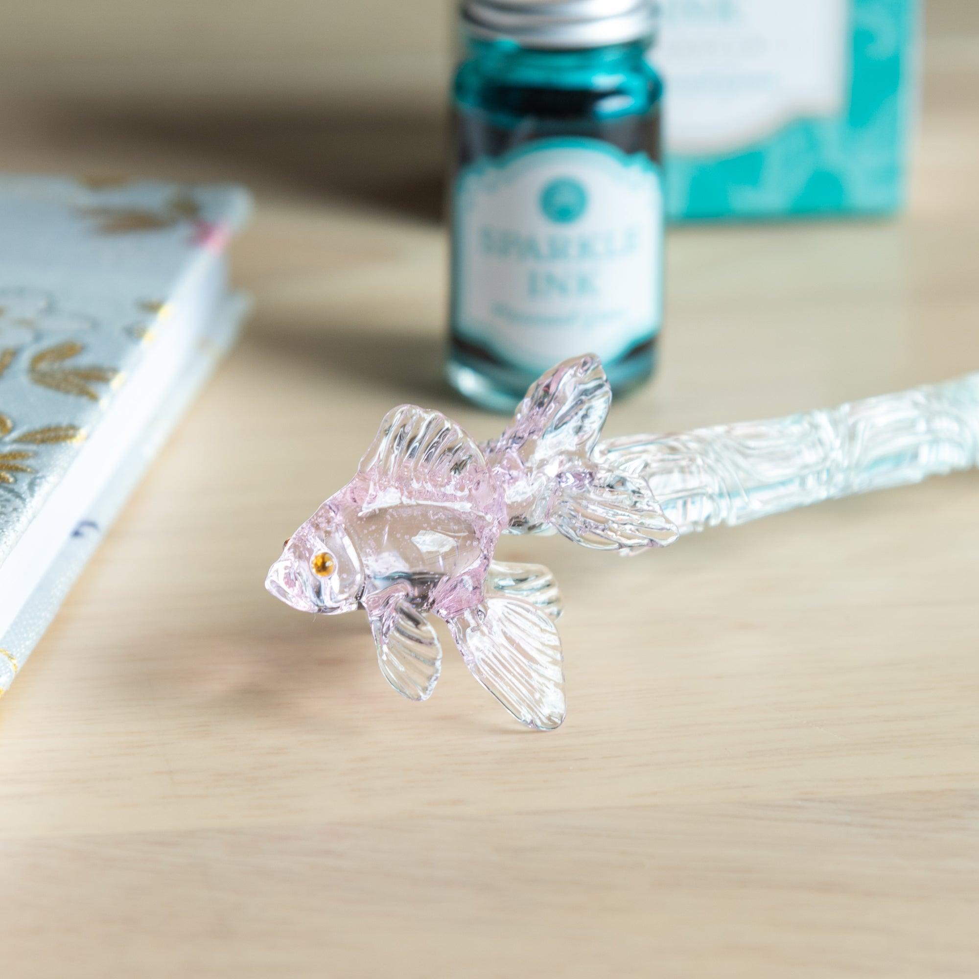 The pink goldfish glass pen held up against a blurry green outdoor background, showcasing the fine craftsmanship and lifelike fish details.