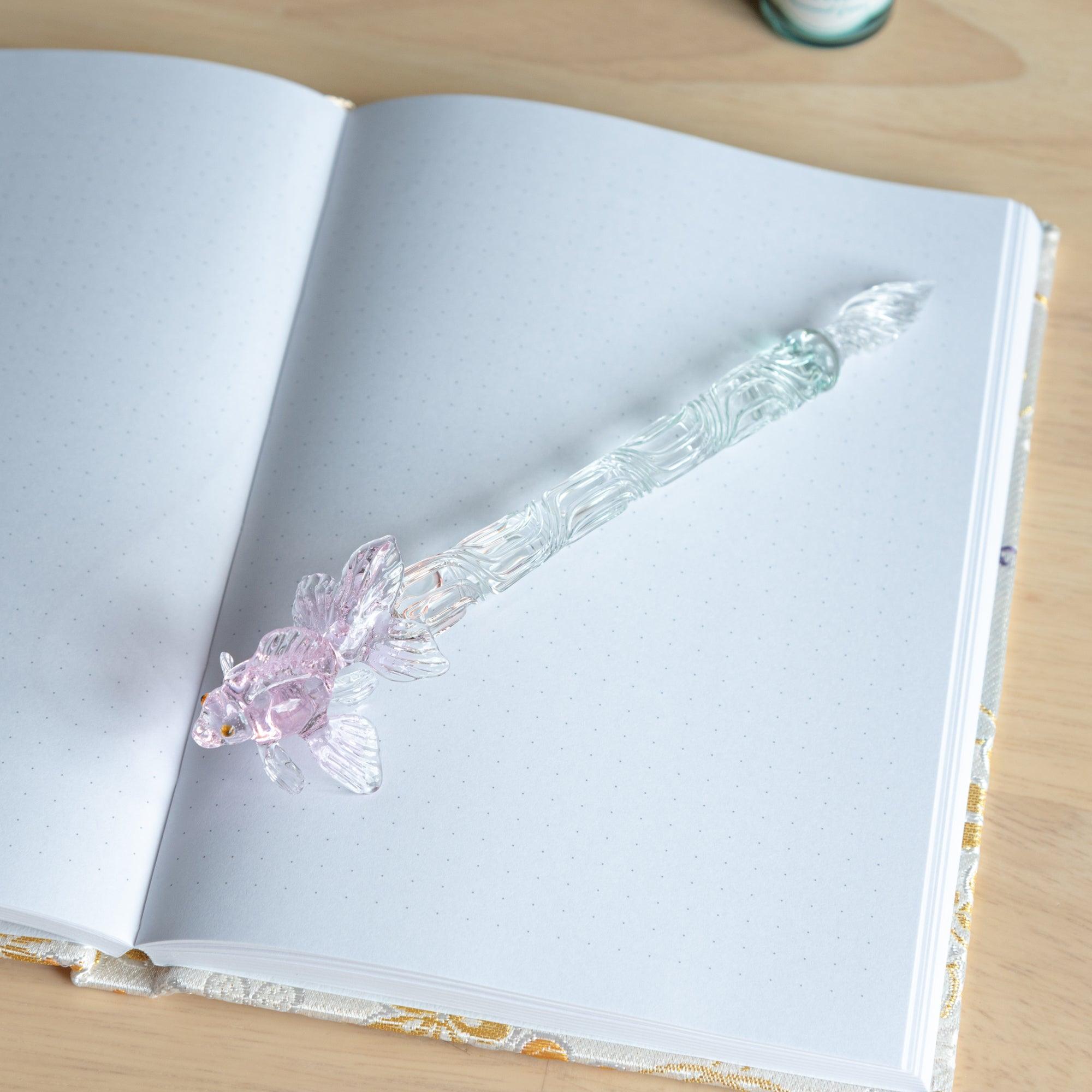 A full view of the pink goldfish glass pen placed on a dotted notebook, highlighting the detailed fish design and swirling glasswork along the pen's shaft.