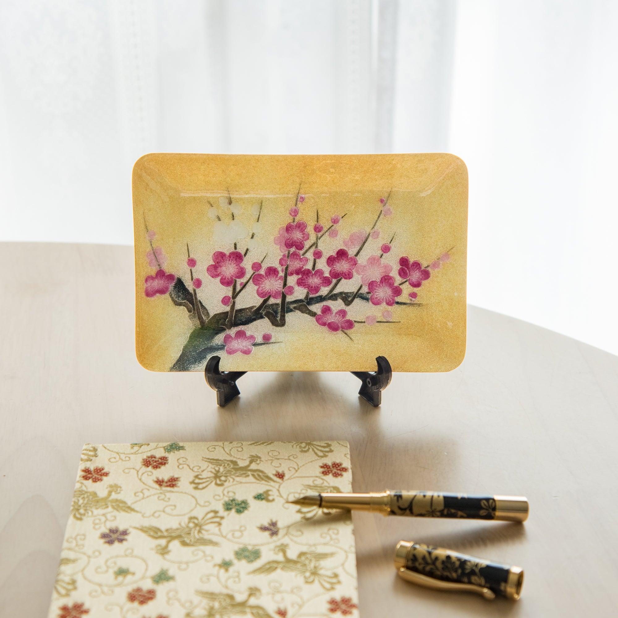 A tray with a plum blossom motif displayed on a stand, paired with a matching patterned notebook and a fountain pen, highlighting Japanese artistry.  