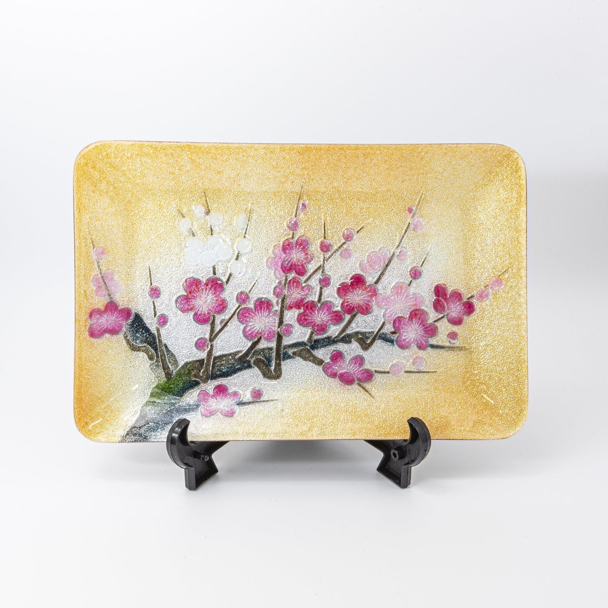 A decorative tray with pink and white plum blossoms on a golden background, displayed upright on a black stand, showcasing Japanese artistry.  