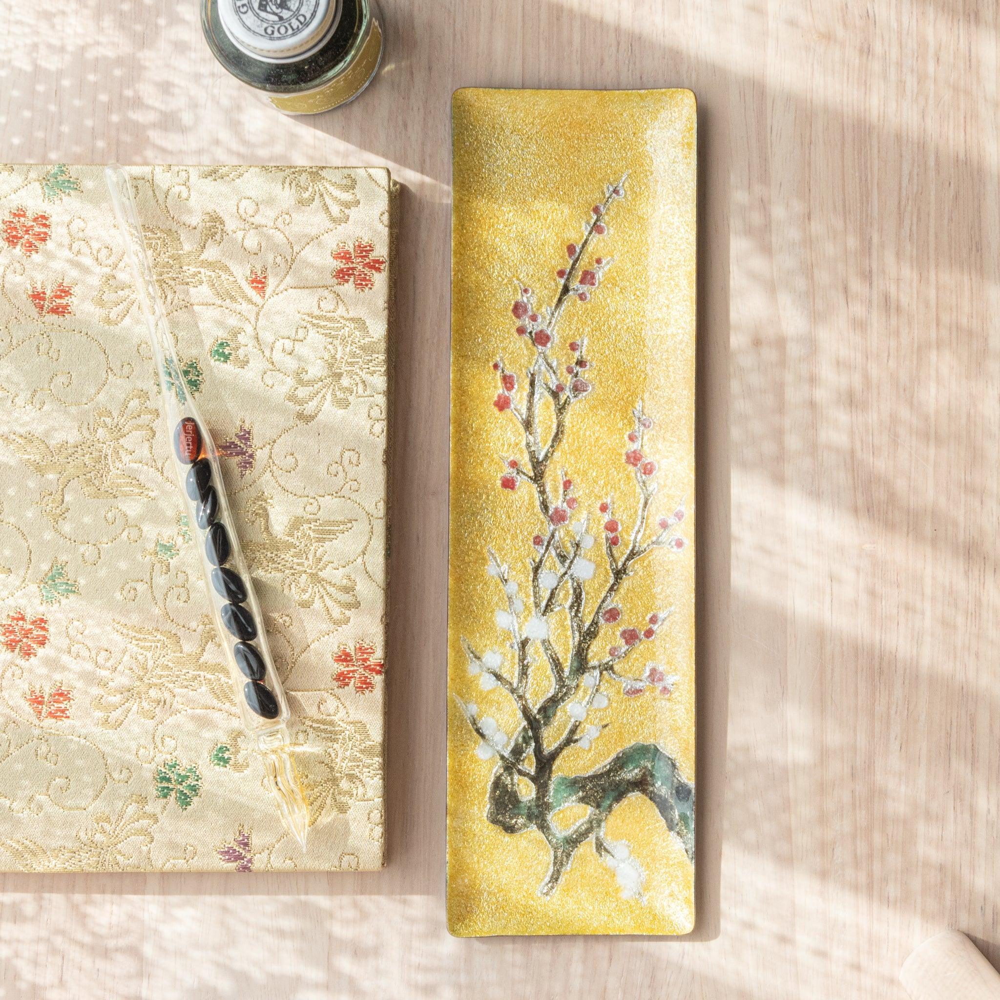 Gold rectangular tray with plum blossom design in white and red, next to a patterned fabric and glass pen, on a light wooden surface.