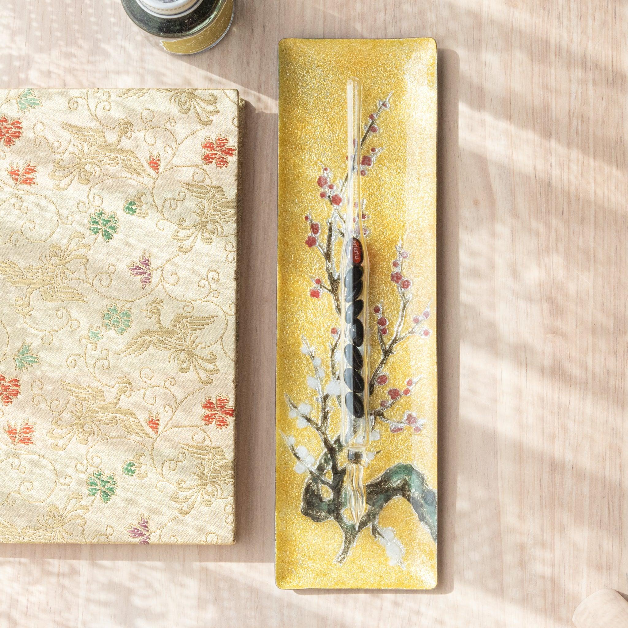 Gold tray with plum blossom motif, featuring a glass pen resting on it, next to a patterned notebook on a wooden background.